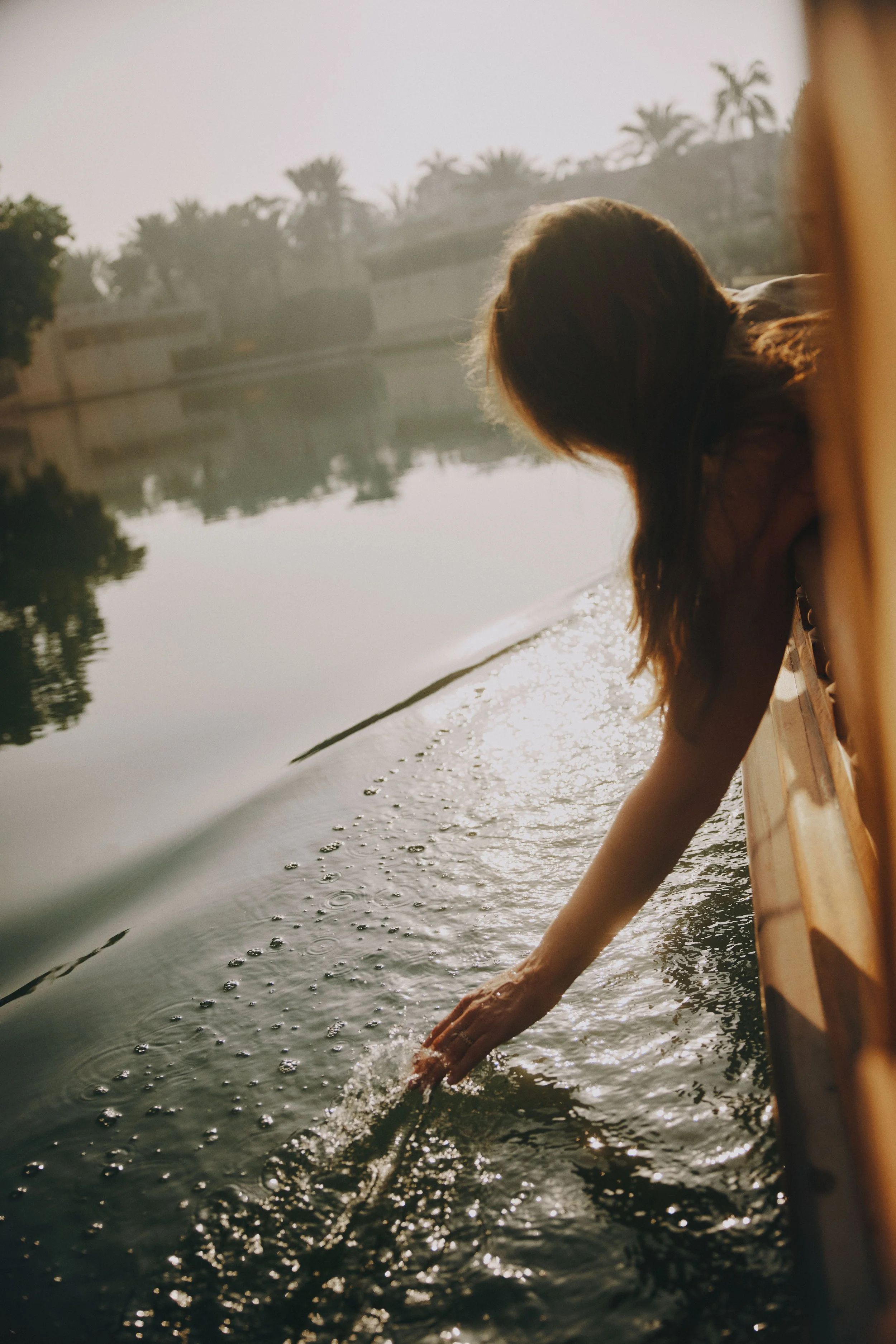 A person with long hair leaning over the edge of a boat, reaching into the water with their hand, during sunset.
