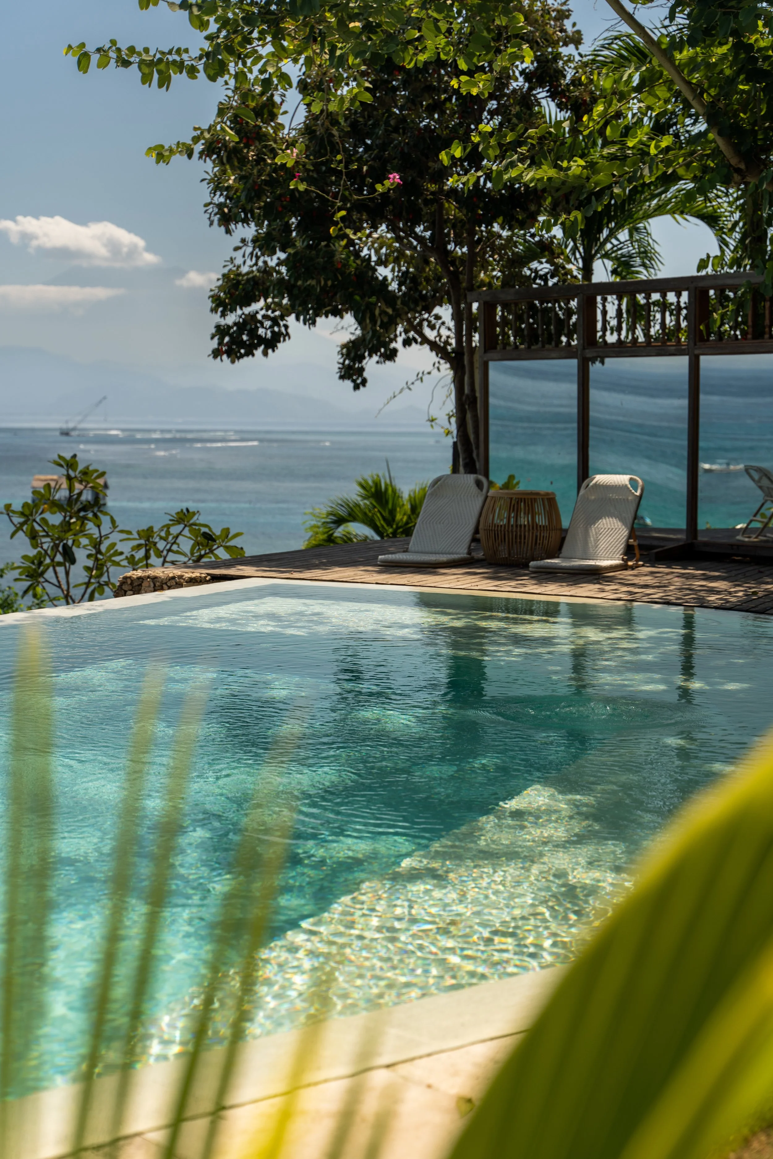 A swimming pool with clear water reflecting the beach and ocean in the background, surrounded by a wooden deck and tropical plants, with lounge chairs and a wicker side table.