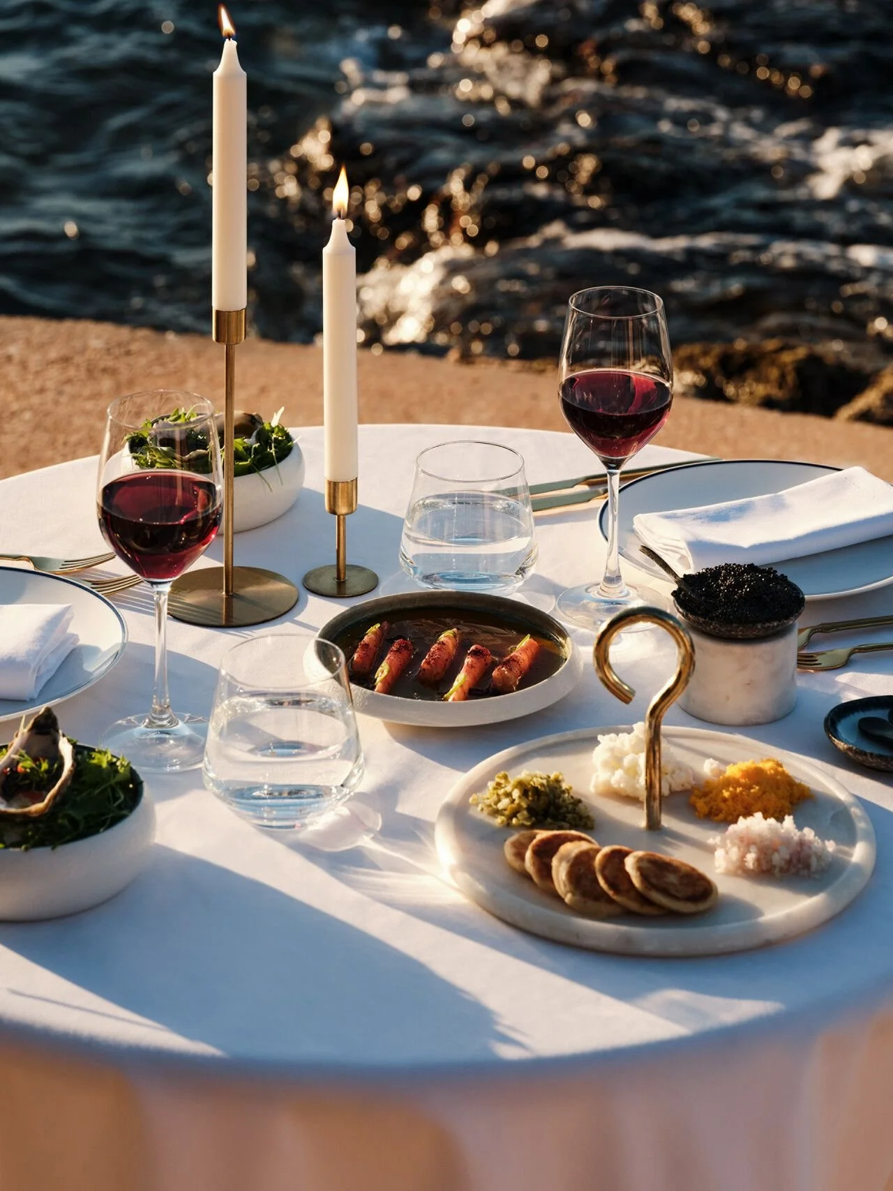 Elegant outdoor dinner table set by the water with white tablecloth, lit candles, red wine, and a variety of appetizers including meats, salads, and breads.