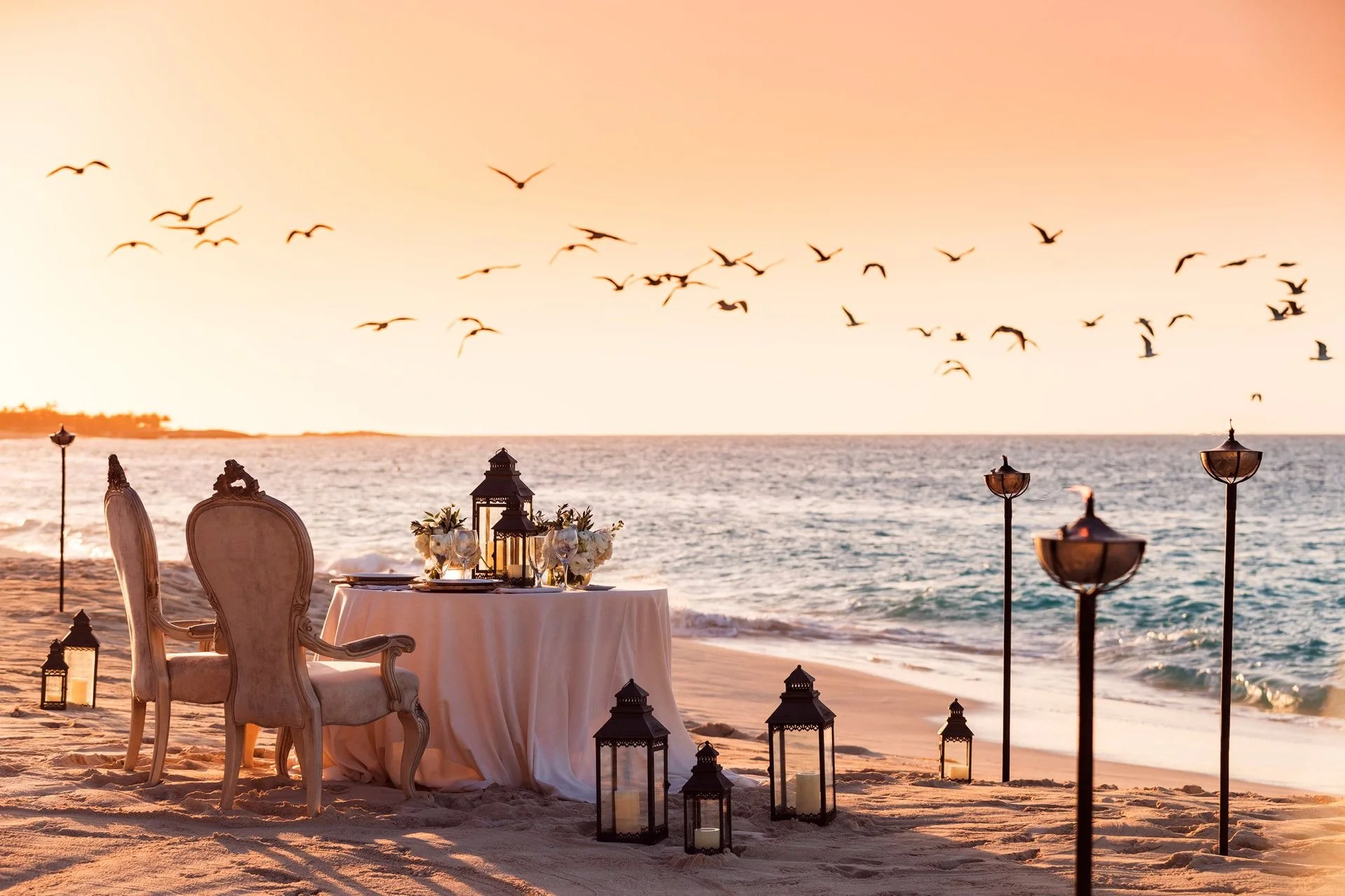 A decorated beach setup for a romantic or elegant event at sunset, with chairs, a table with flowers, lanterns, and a view of the ocean and seagulls flying in the sky.