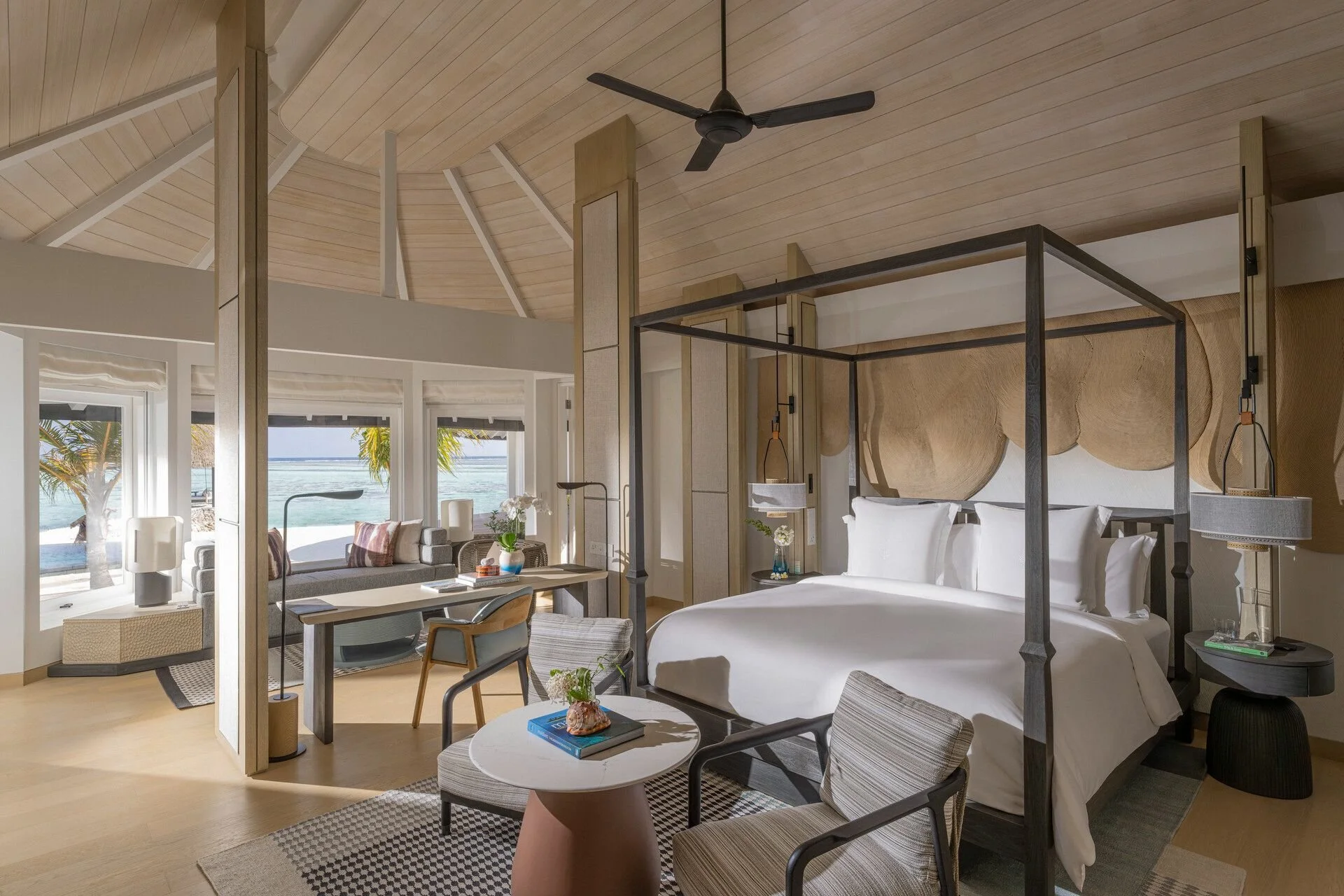 Interior of a modern beachfront hotel room with a canopy bed, sitting area, and ocean view through large windows, featuring wooden ceiling, ceiling fan, and stylish decor.