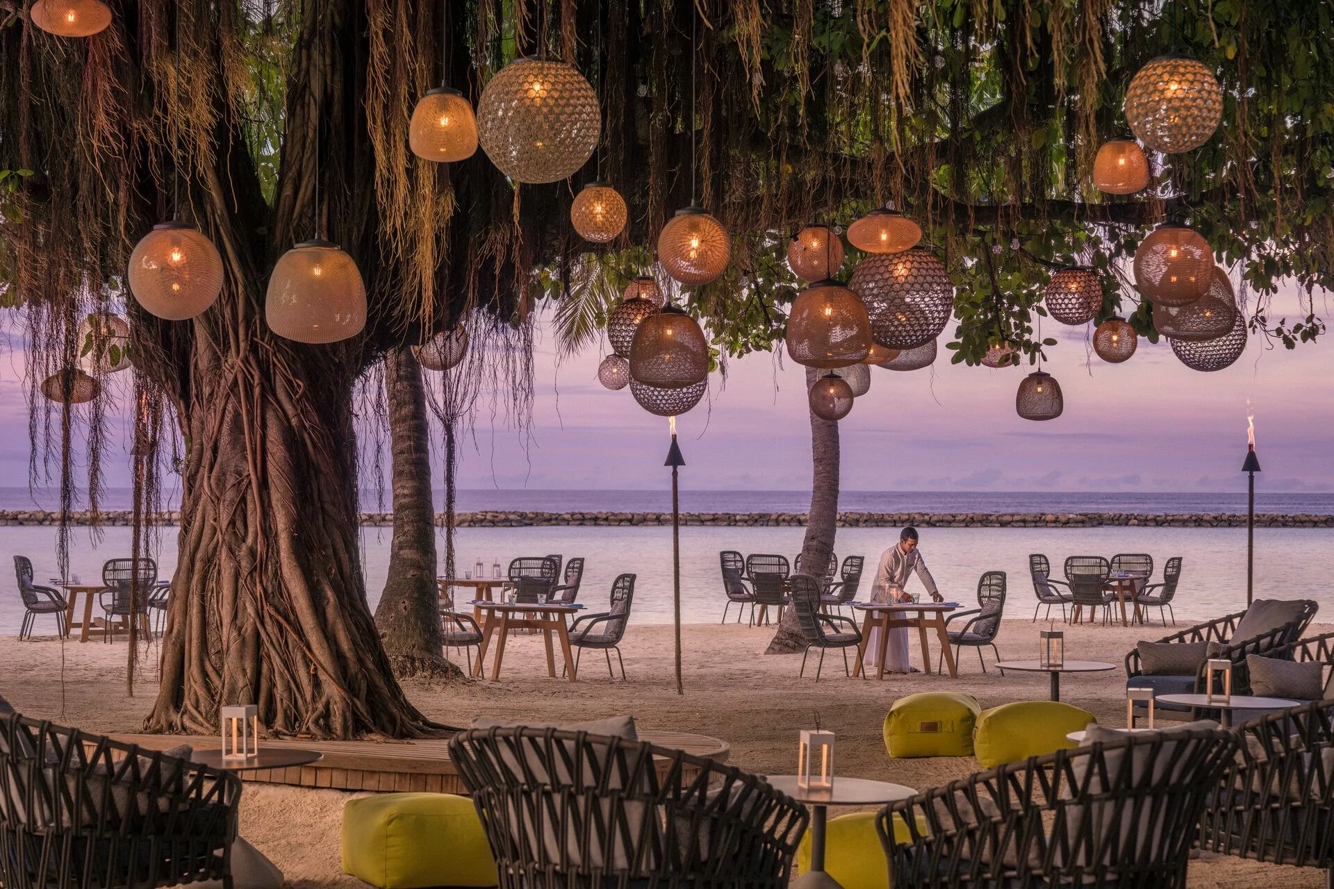 Beachside restaurant with hanging lanterns, outdoor tables, chairs, potted plants, and a man setting a table near the ocean at sunset.