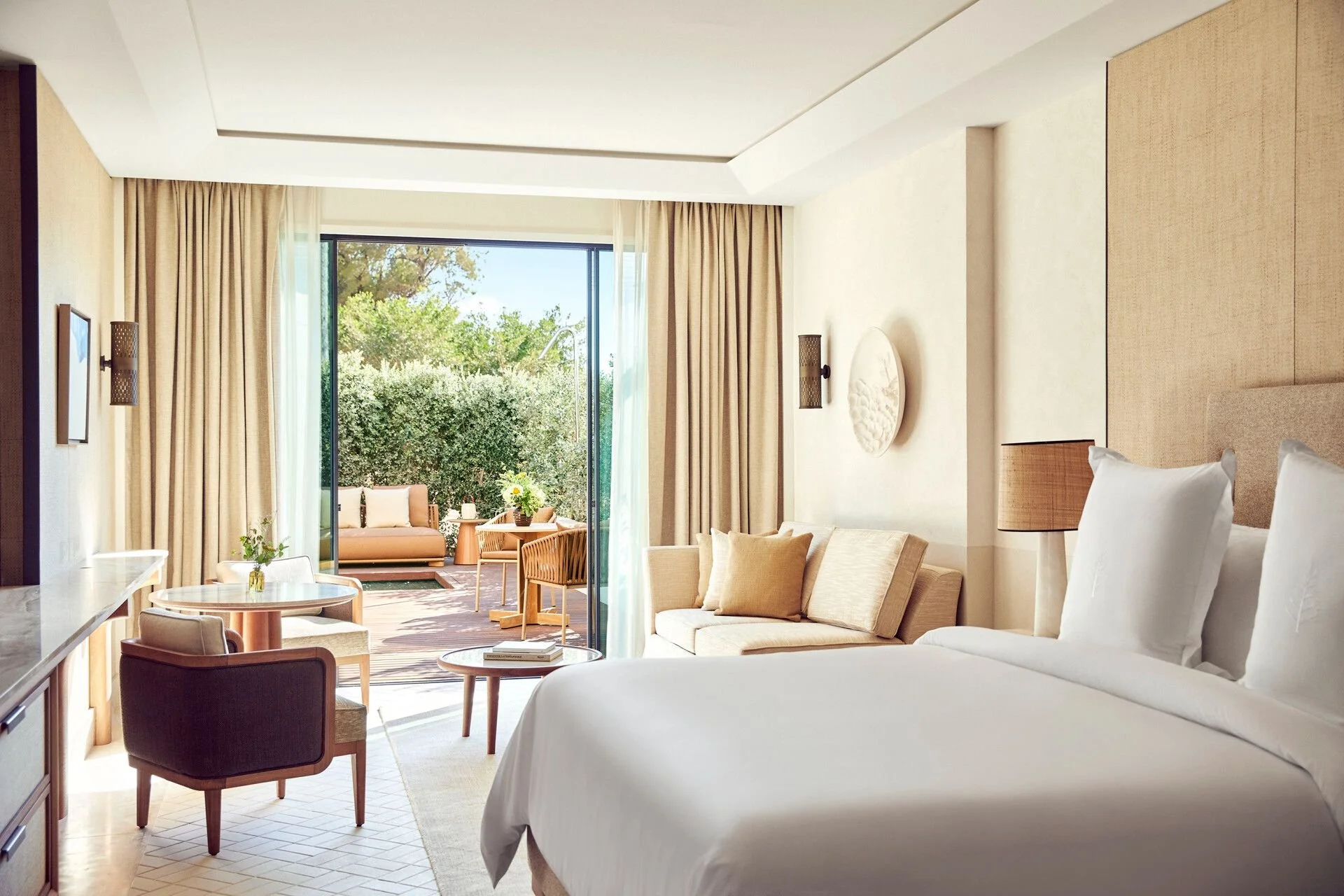 Interior of a modern hotel room with a bed, sitting area, and balcony with outdoor seating, greenery, and trees outside.
