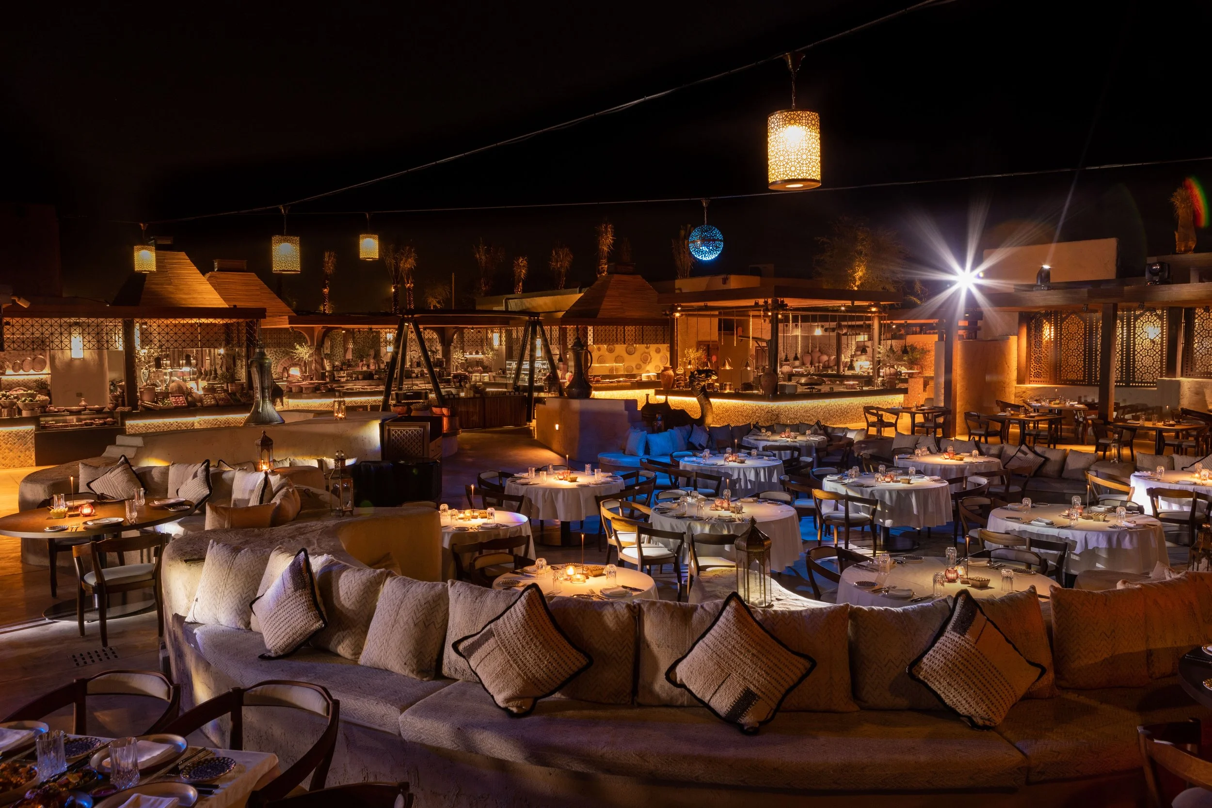A nighttime view of an outdoor restaurant or lounge area with round tables, plush seating, and decorative hanging lanterns and spherical lights, illuminated with warm lighting.