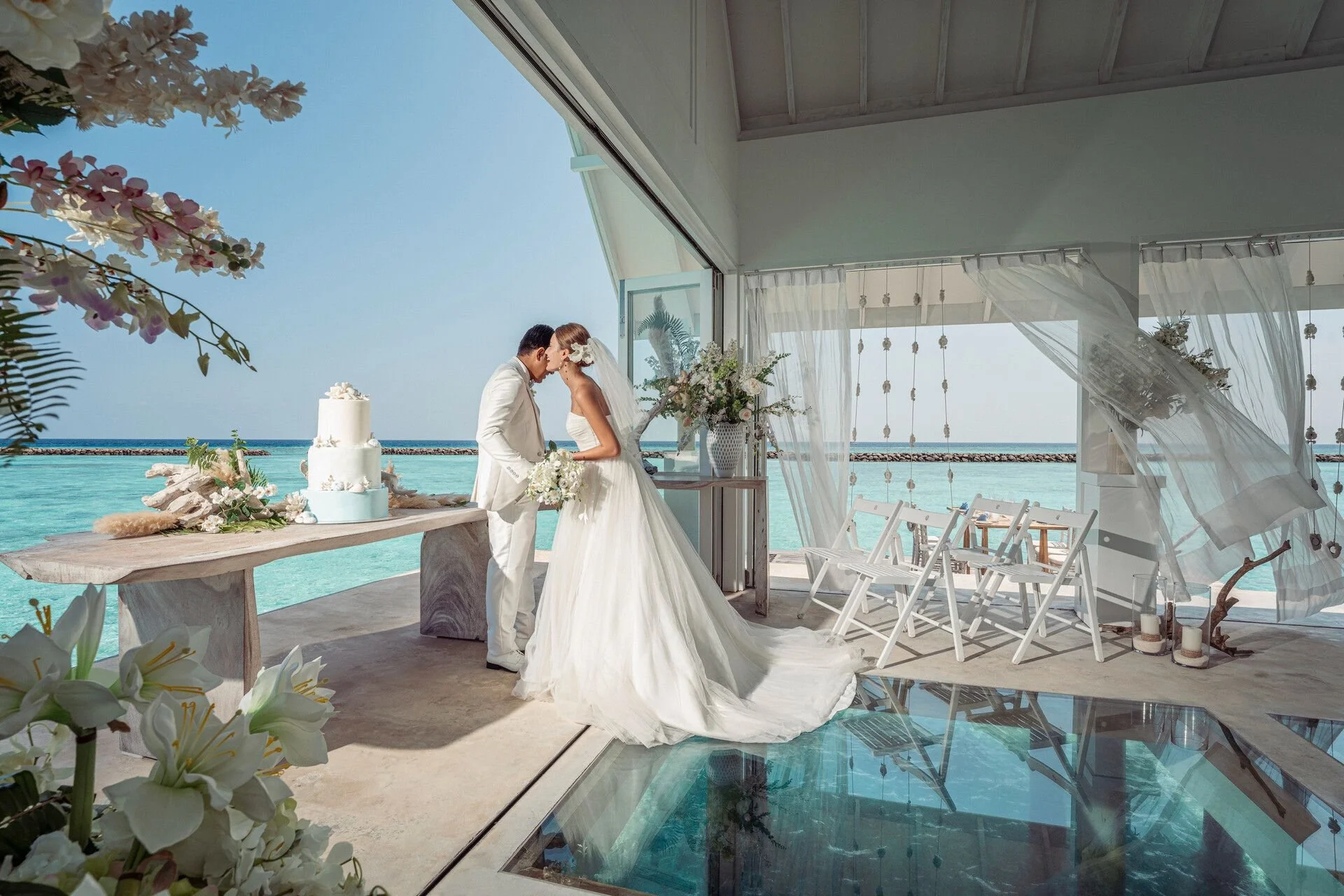 A bride and groom share a kiss during their wedding ceremony on a beachside pavilion with sheer curtains, white chairs, and floral decorations, overlooking the ocean.