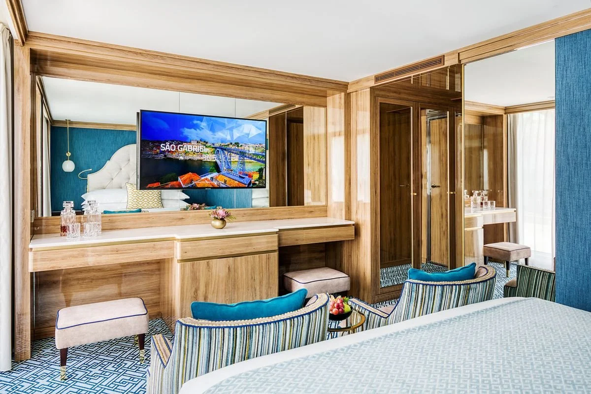 Hotel room with a large mounted flat-screen TV, blue and striped armchairs, a bed with light-colored bedding, and wood-paneled walls and furniture.