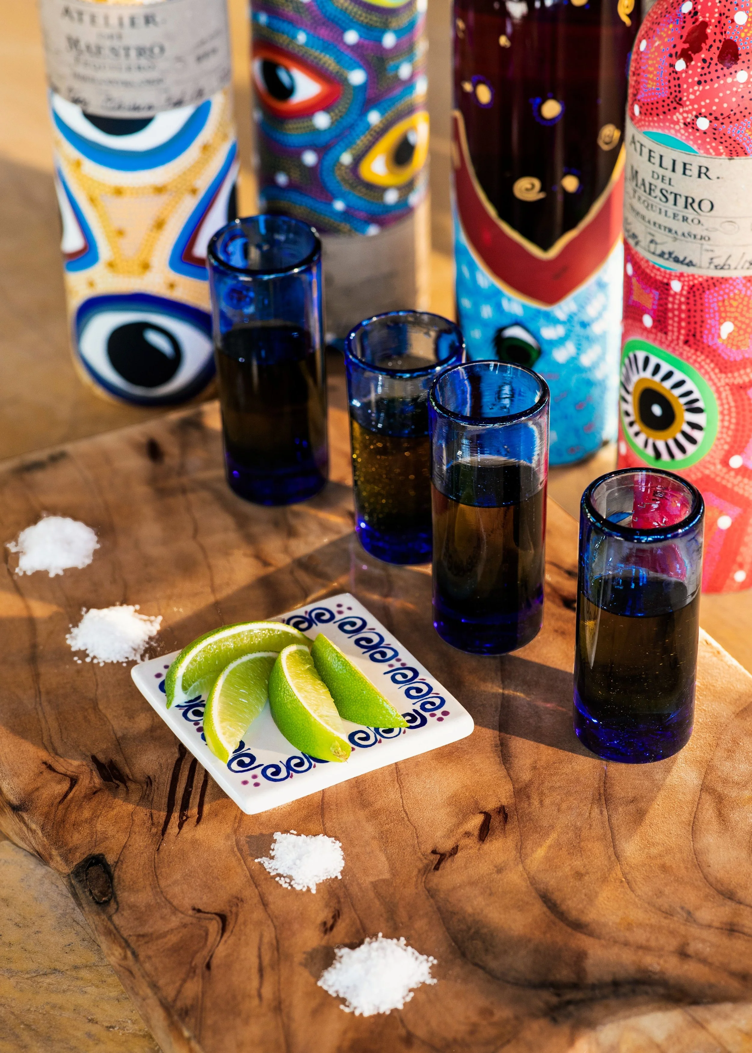 Colorful bottles with artistic designs, shot glasses filled with dark liquid, a small plate of lime wedges, and scattered salt on a rustic wooden table.