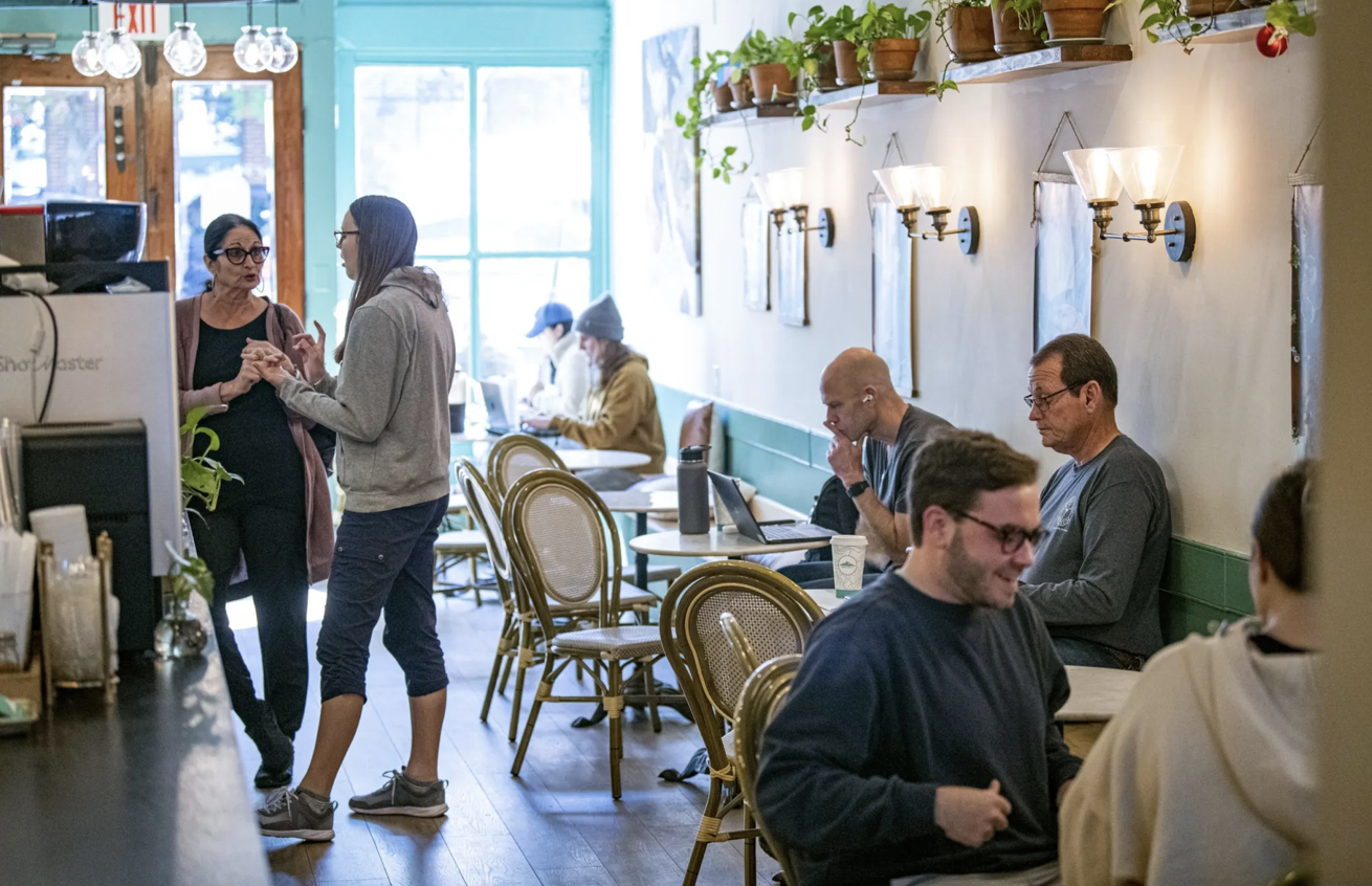 People inside a cozy cafe, engaging in conversations and working on laptops, with a vibrant turquoise wall, potted plants, and warm lighting.