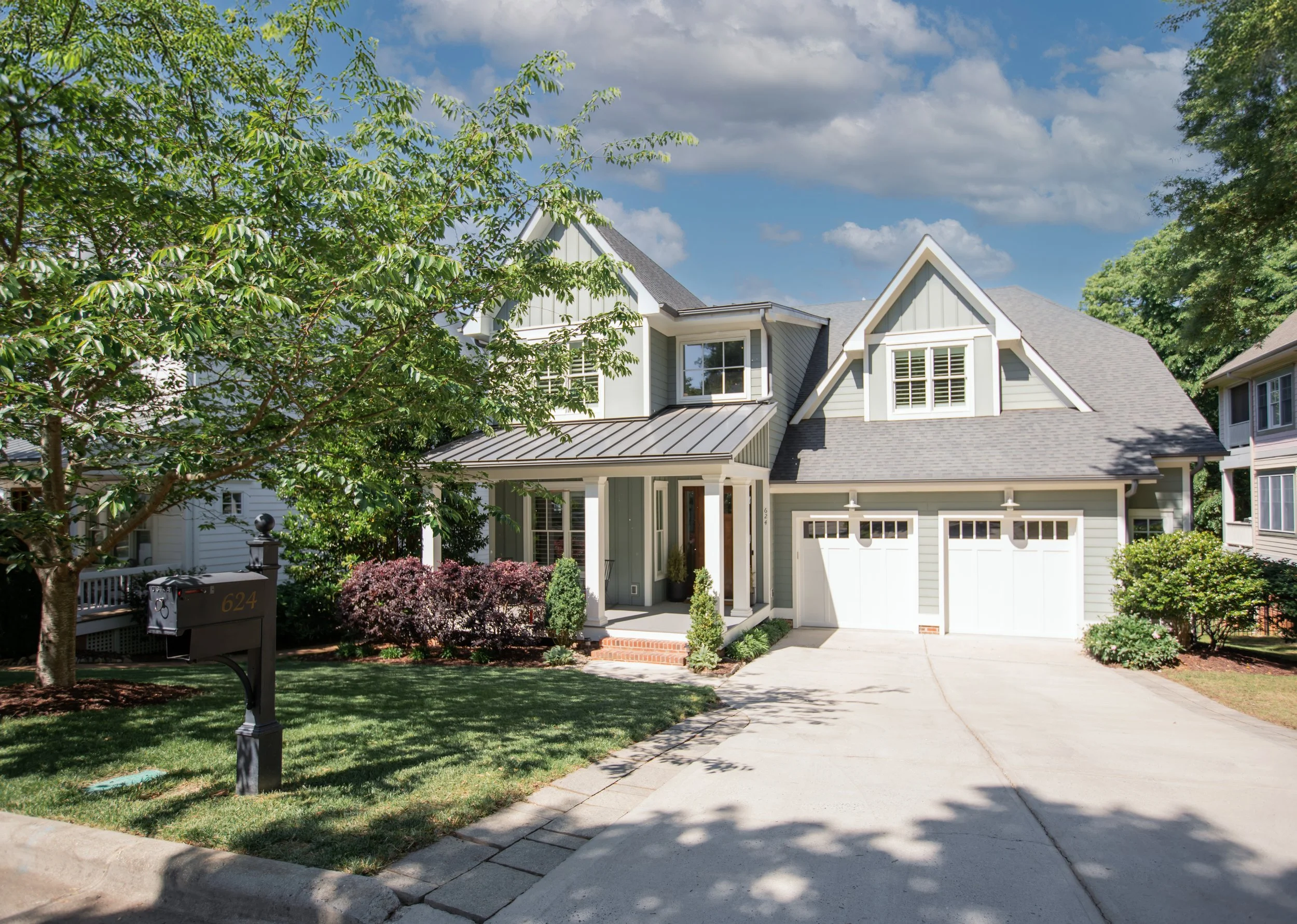A two-story suburban house with gray siding and white trim, featuring a front porch, a garage, and a neatly landscaped yard with trees and shrubs on a sunny day.