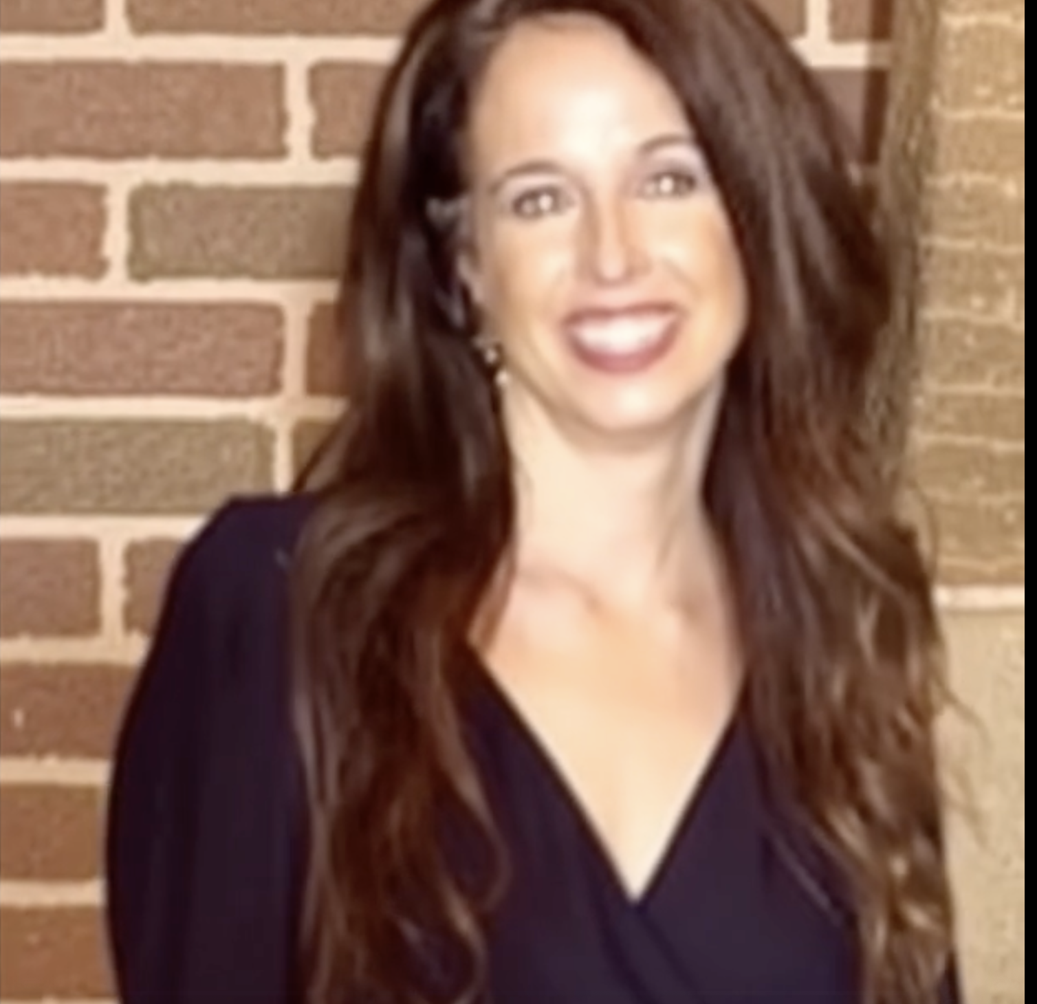 A woman with long, wavy brown hair smiling in front of a brick wall.