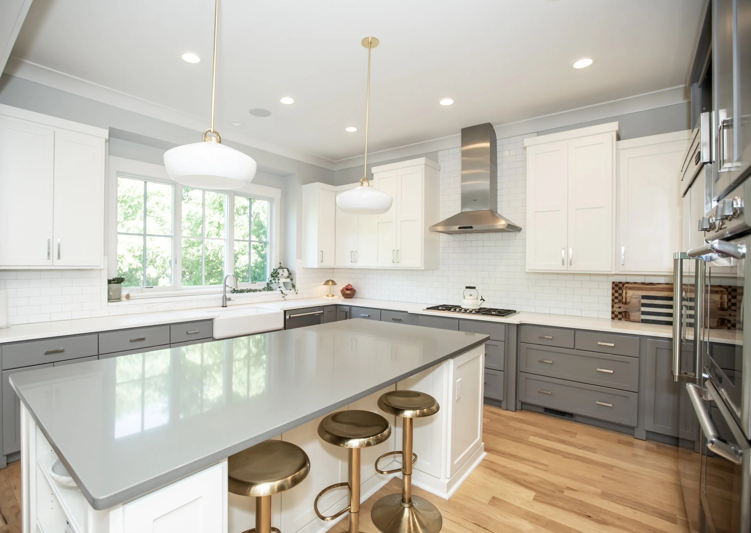 Modern kitchen with white and gray cabinets, large window letting in natural light, island with gray countertop, and bar stools with brass bases.