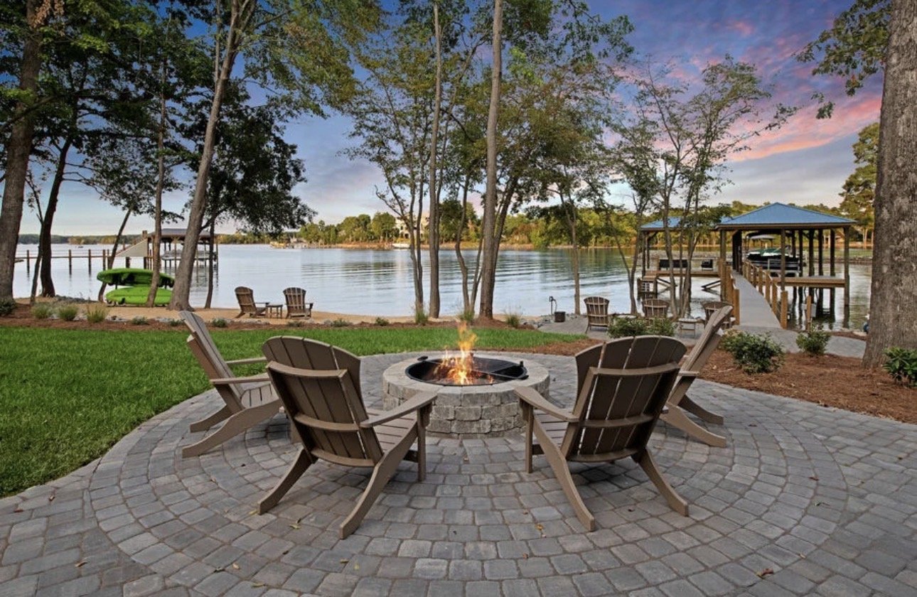 A lakeside scene at sunset with a fire pit surrounded by six chairs on a stone patio, trees, a dock with boats and a covered pavilion, and a colorful sky.