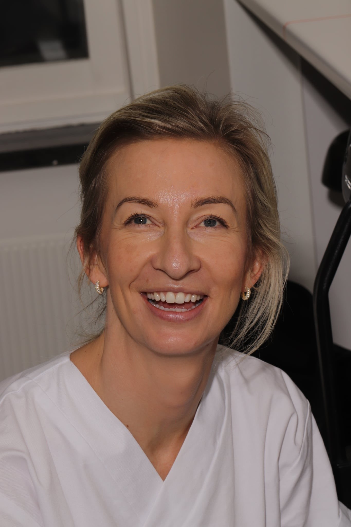 Smilende kvinne med kort hår og ballerinaer, iført hvit uniform, i et rom som ser ut som en klinikksal.