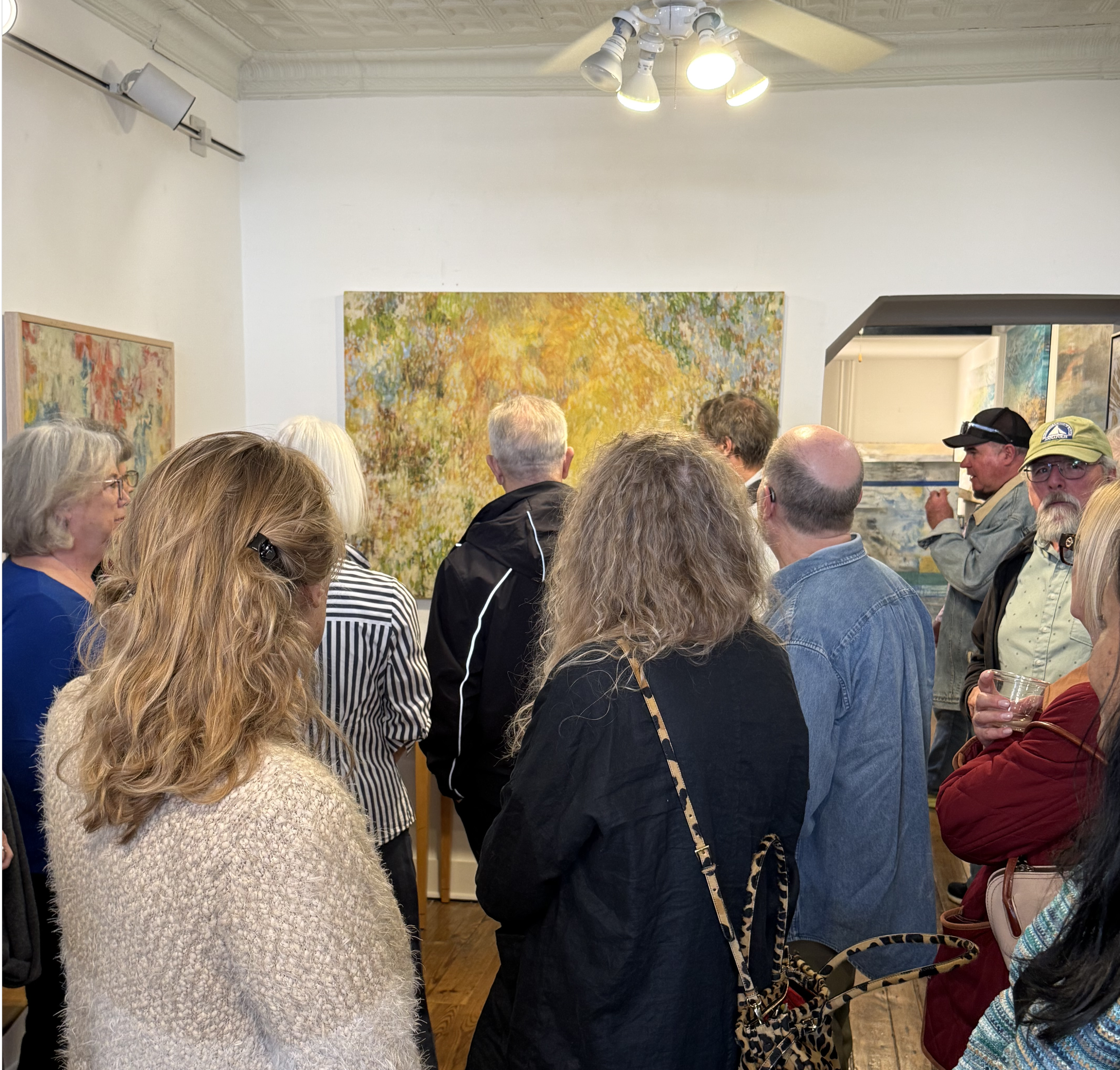 A crowd of people at an art gallery or exhibition, viewing colorful paintings on the walls. Some individuals are engaged in conversation, while others are observing the artwork.