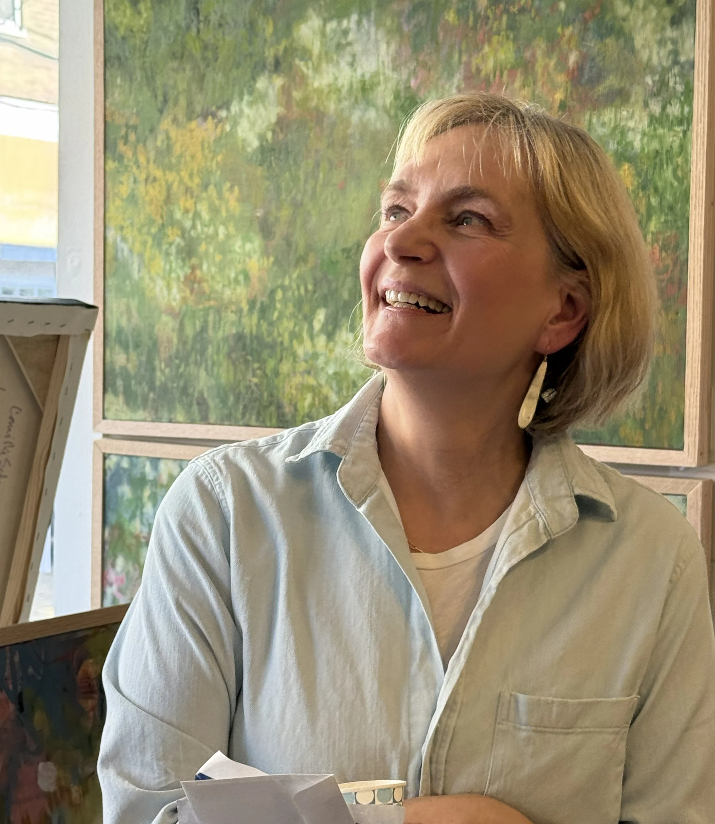 An older woman with blonde hair, smiling and wearing a beige button-up shirt, sitting at a table in front of a background of framed colorful paintings of nature scenes.