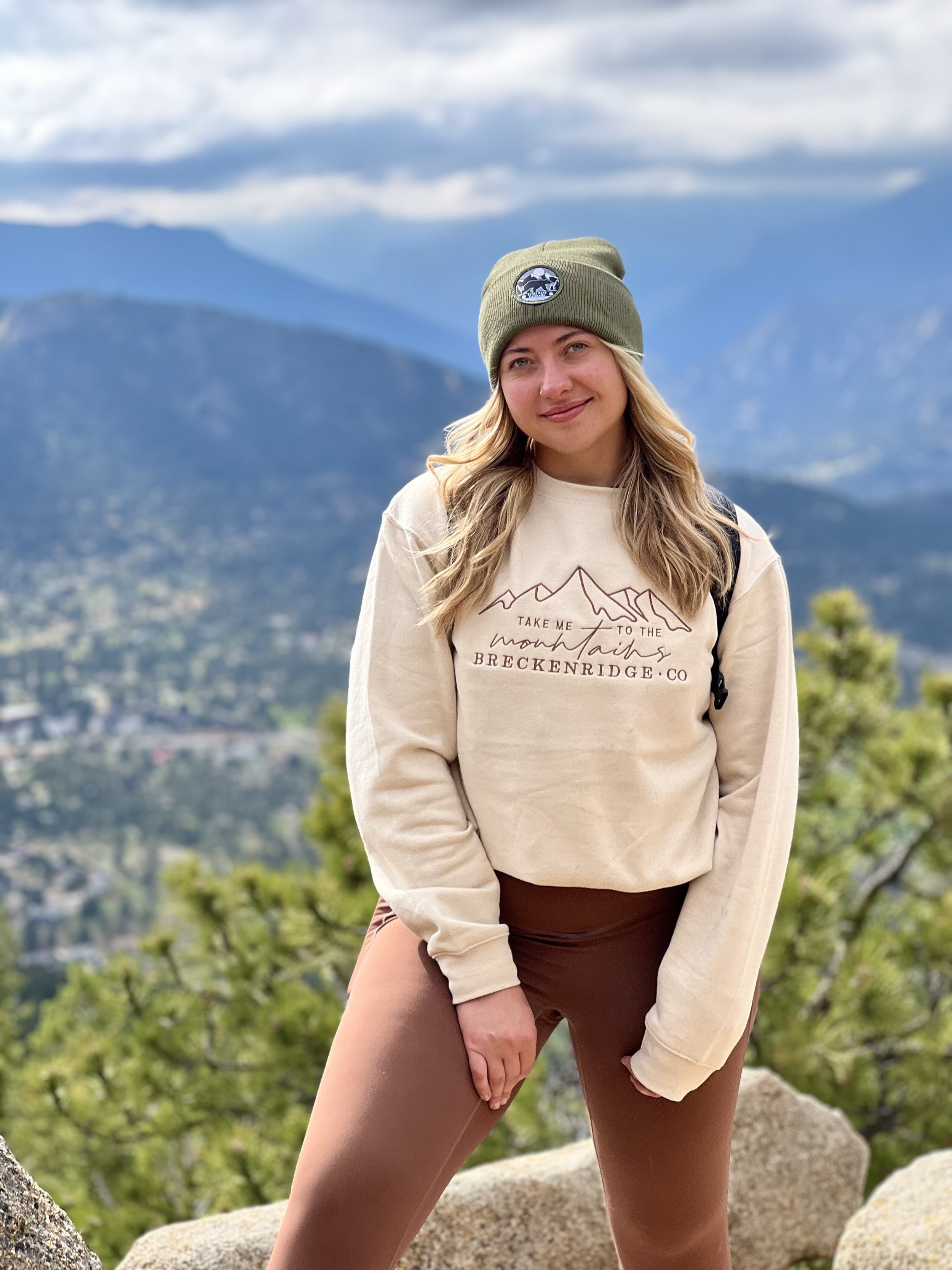 Blonde woman wearing a Take Me to the Mountains in Breckenridge, CO cream colored sweatshirt, brown leggings, and a green hat with the mountains of Estes Park, CO in the background.