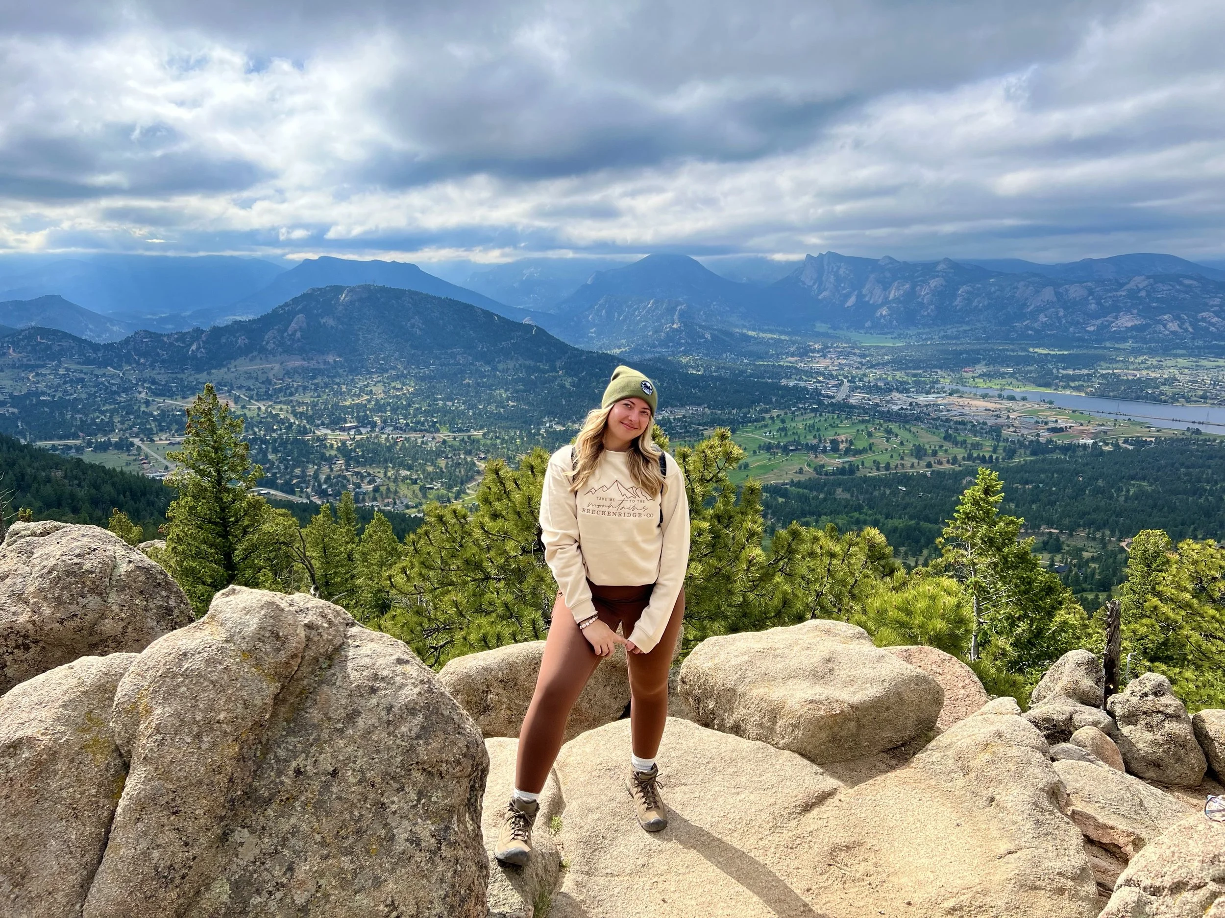 Blonde woman wearing a "Take me to the mountains of Breckenridge, CO" cream colored sweatshirt, brown leggings and a green hat with the mountains of Estes Park, CO in the background.