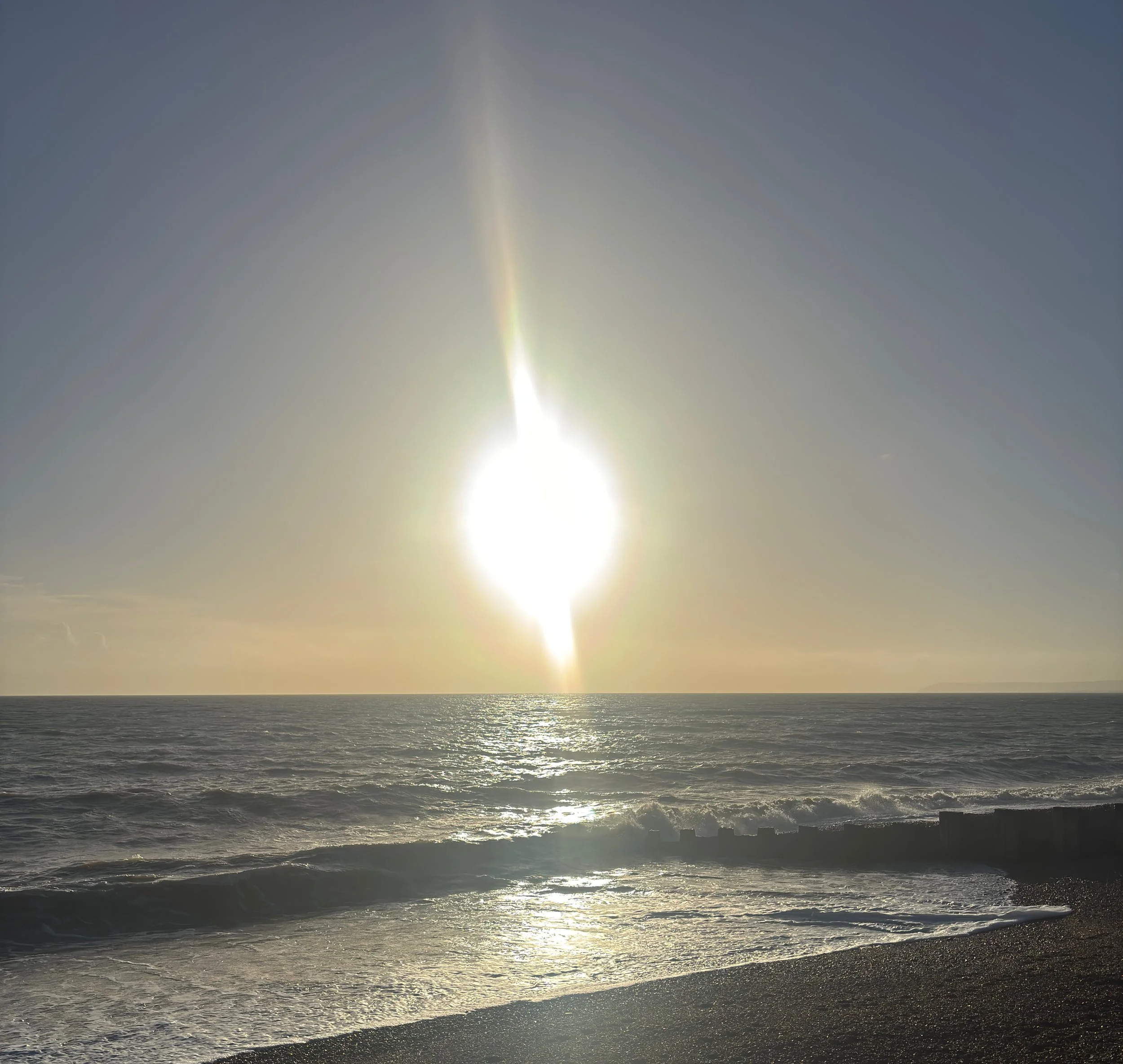 View of the ocean with gentle waves and the sun low on the horizon, creating a bright glare on the water.
