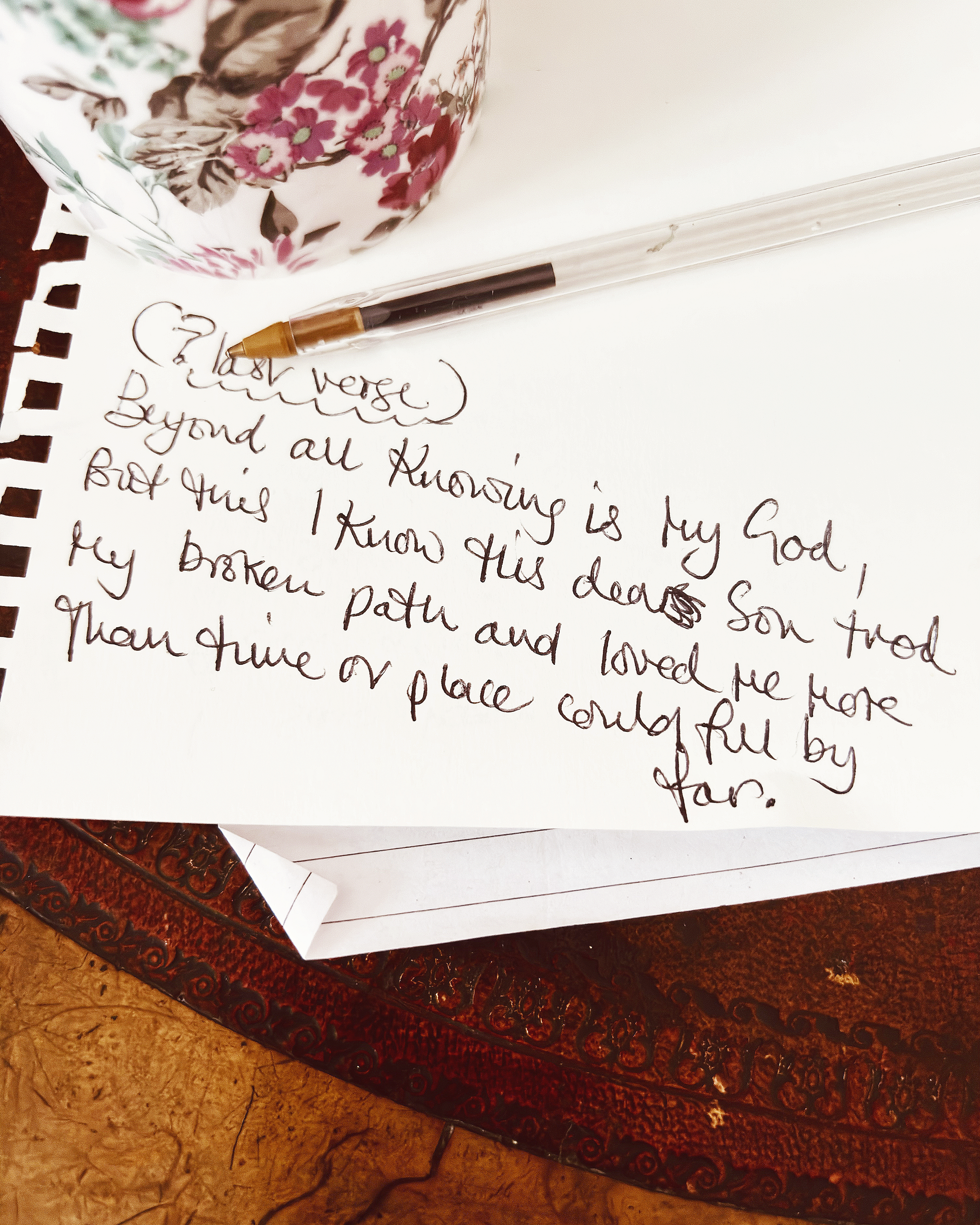 Close-up of handwritten song lyric on note paper, partially visible, with a decorative floral-patterned mug in the background, placed on an old dark wooden table.