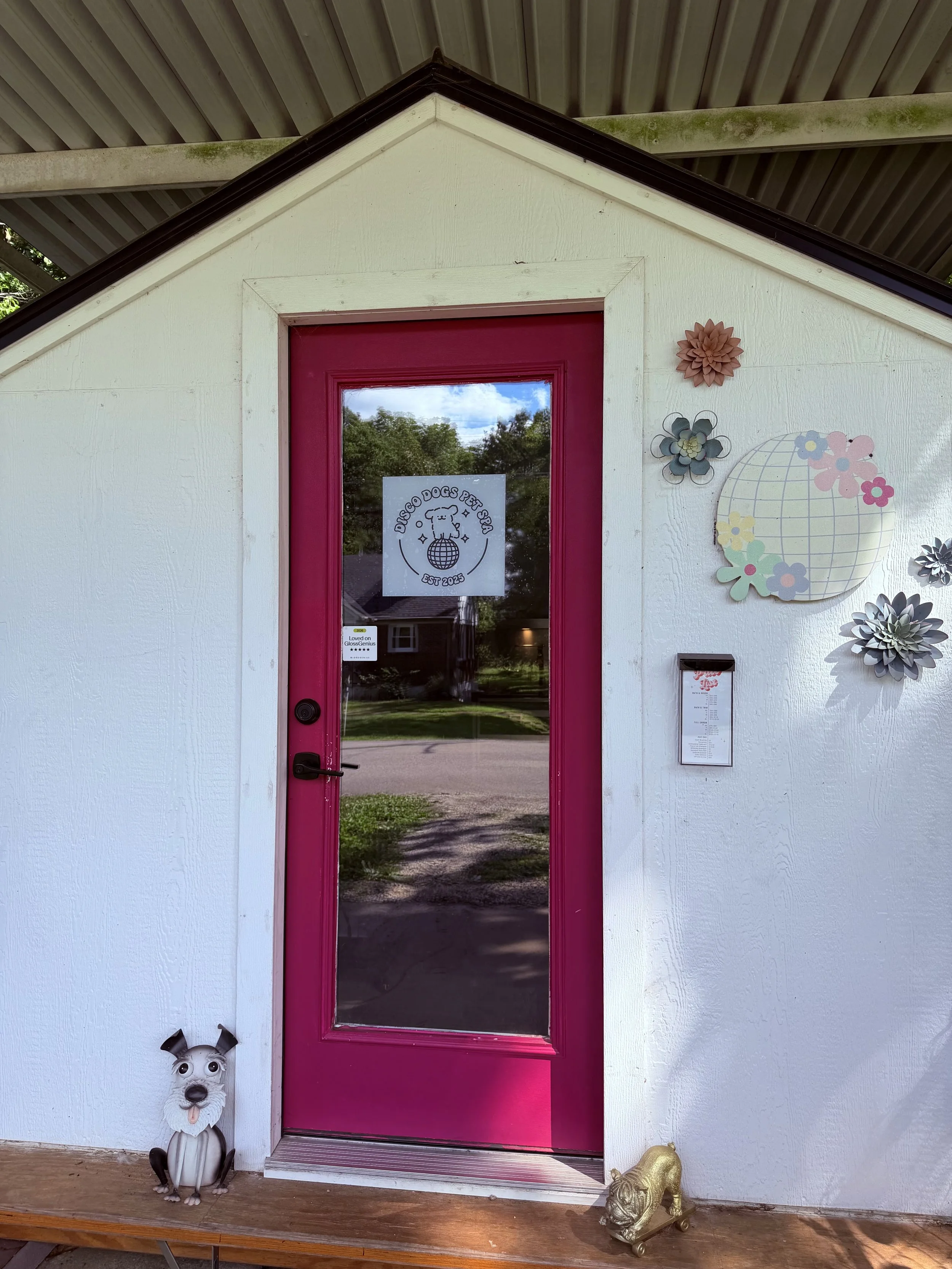 White building with pink door, decorative metal flower wall art, and dog figure at the bottom left corner.