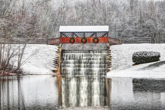 A small covered dam with a roof, three orange life rings attached, and a waterfall flowing into a pond, with snow-covered ground and leafless trees in the background.