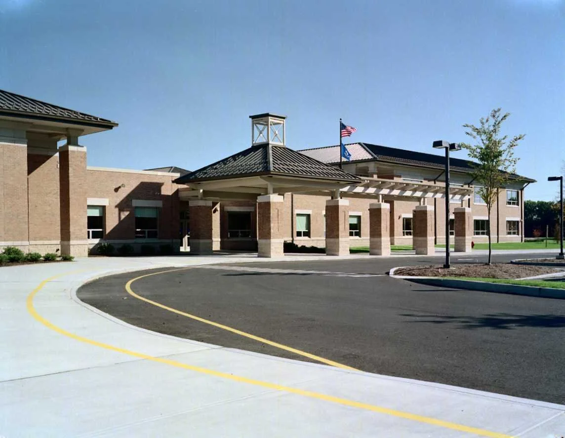Exterior view of a modern brick school building with a covered entrance, American flags, and a parking lot with yellow lines.
