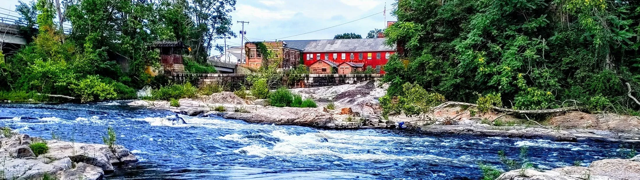 A river flowing over rocks surrounded by greenery with red brick buildings and utility poles in the background