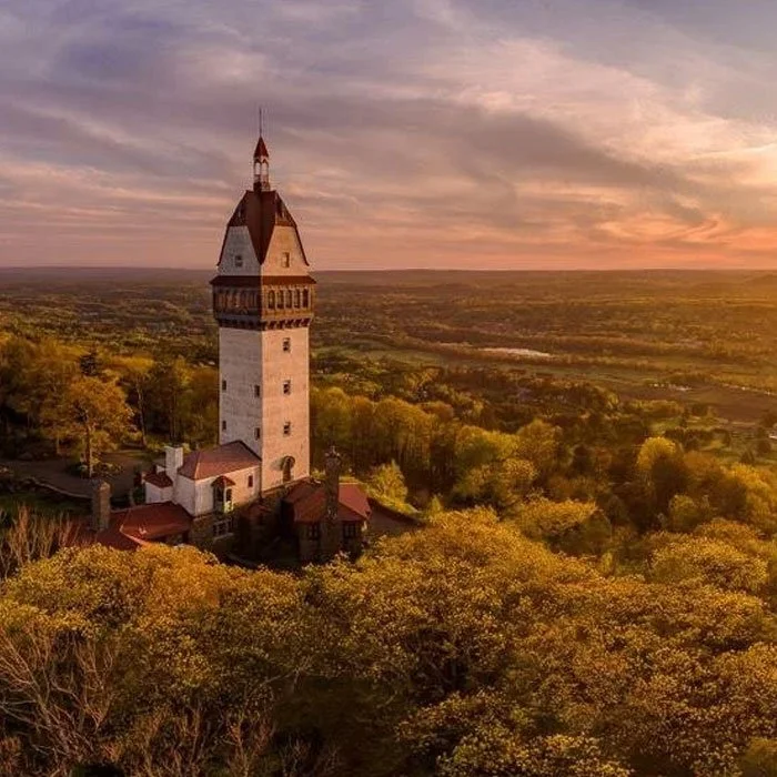 A tall historic building with a pointed roof and a tower, set among autumn trees with a sunset sky in the background.