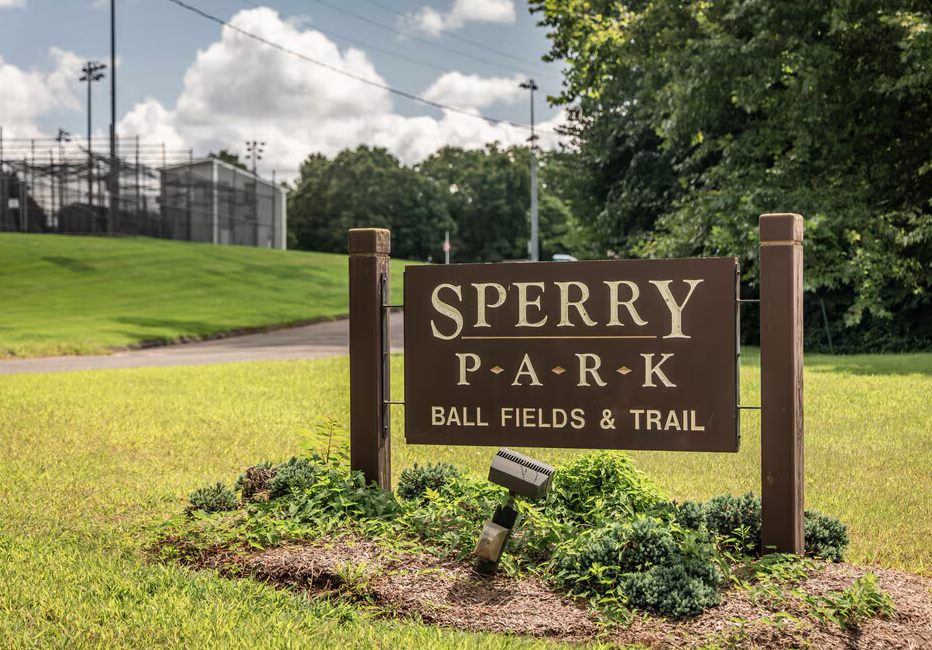 Sign for Sperry Park with ball fields and trail, surrounded by green grass and trees, near a sports field or sports complex.