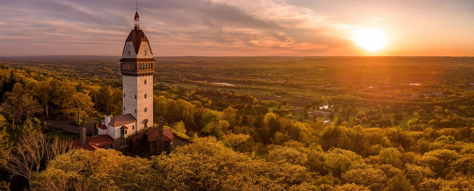 A tall historic tower with a pointed roof surrounded by autumn trees during sunset.