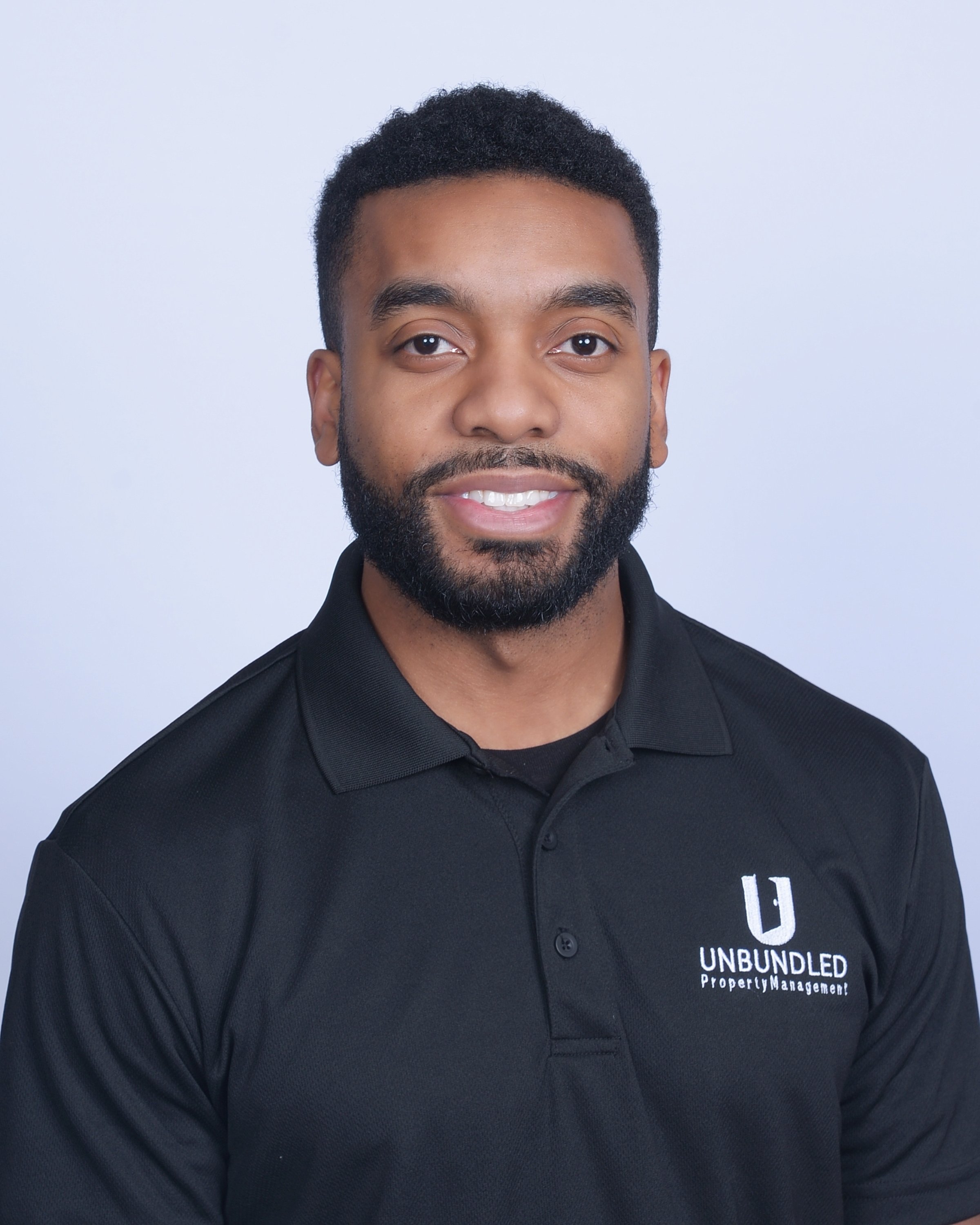 A man smiling wearing a black polo shirt with a logo and text that reads "UNBUNDLED Property Management" on the chest, against a plain light background.