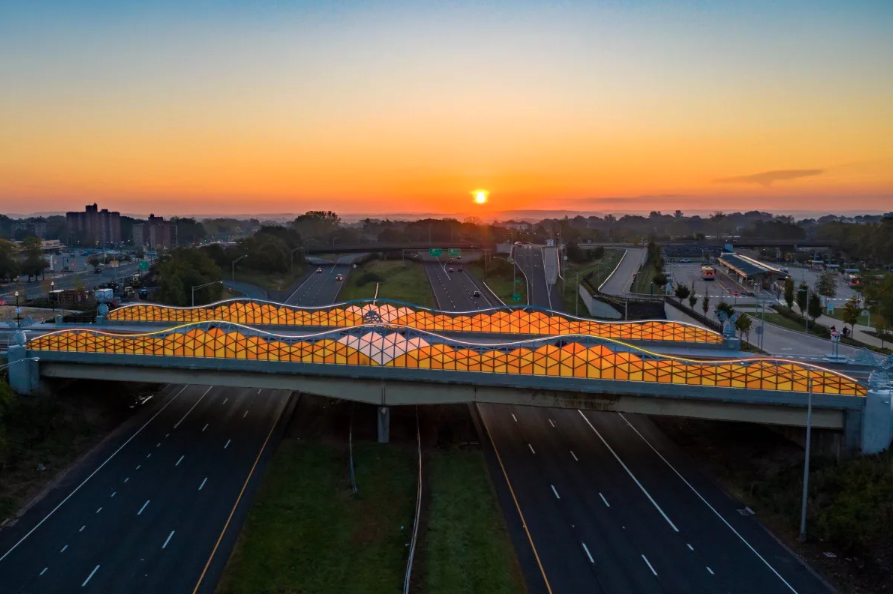 An aerial view of a highway overpass with a colorful, wave-like canopy design at sunset, with trees, buildings, and a parking lot in the background.