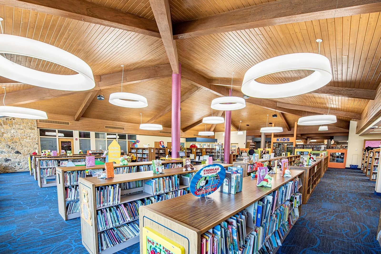 A brightly lit children's section in a library with wooden bookshelves and a high wooden ceiling featuring large, modern circular light fixtures. The shelves are filled with colorful books and children's toys, including stuffed animals. There is a sign that reads 'KIDS' in the background.