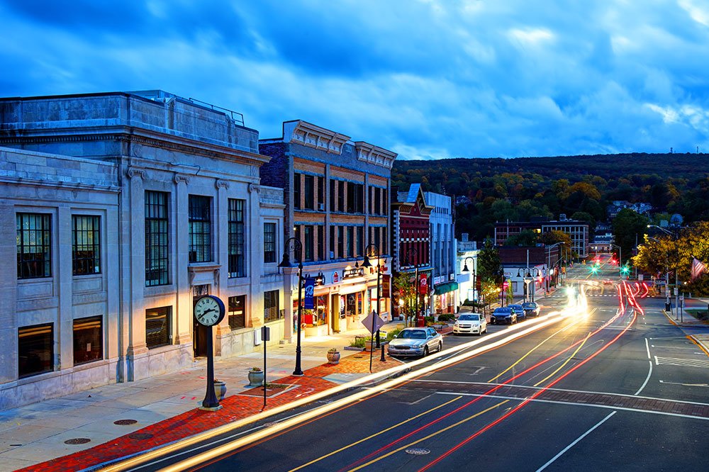 A small town main street at dusk with historic buildings, parked cars, streetlights, and light trails from moving vehicles.