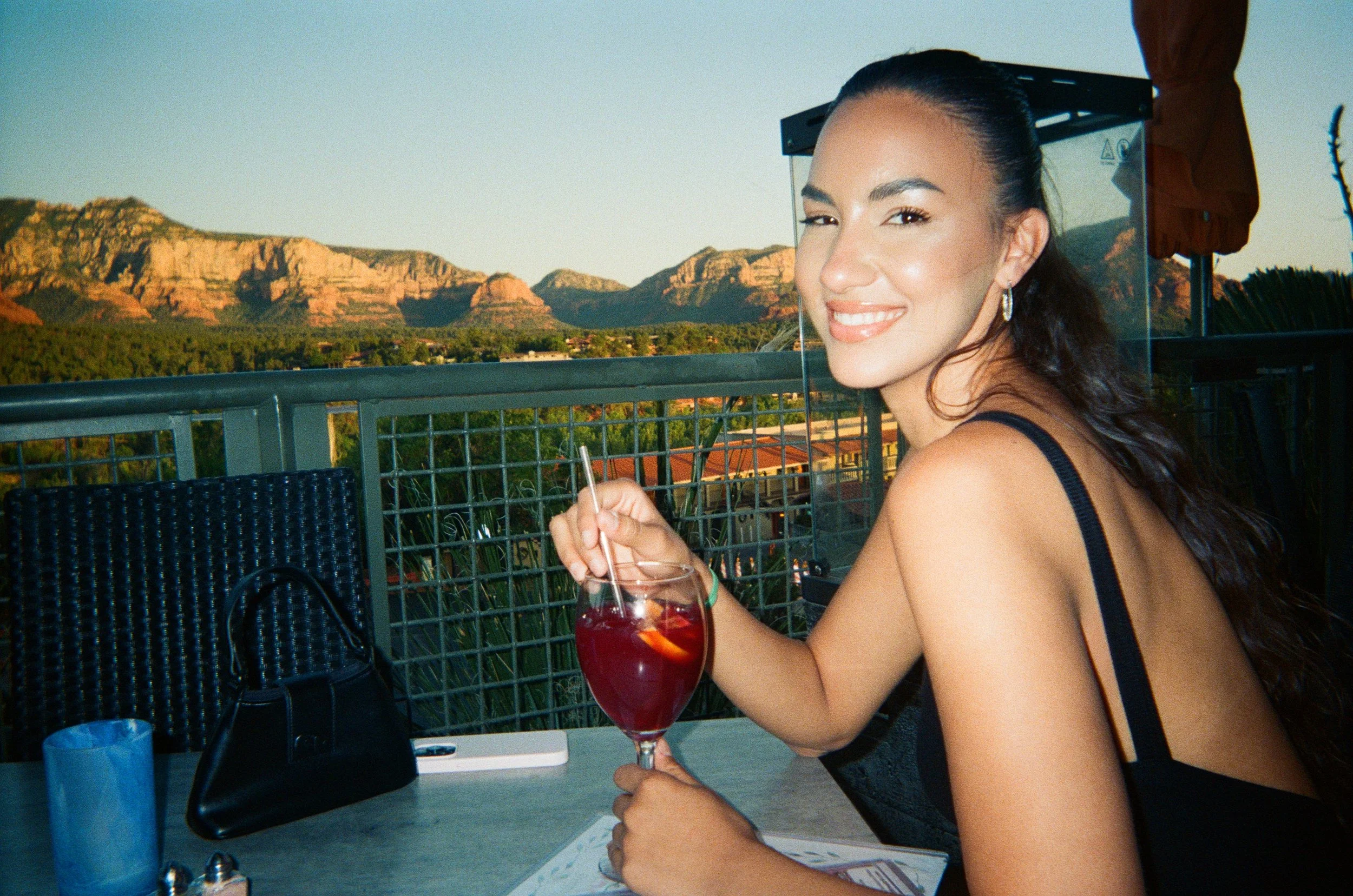 A woman with dark, wavy hair and hoop earrings, smiling and winking, sitting at an outdoor table with a glass of red drink. In the background, there are mountains and a clear sky.