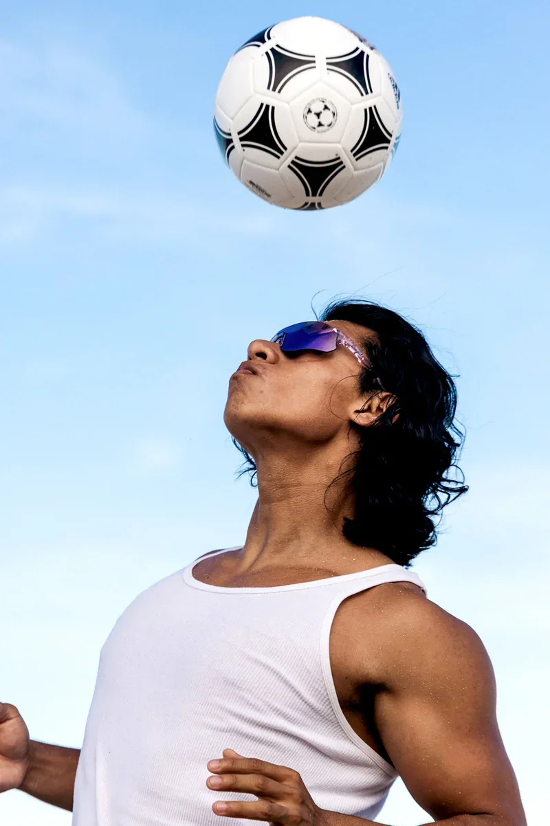 A person with black hair wearing sunglasses and a white tank top, heading a soccer ball against a blue sky.