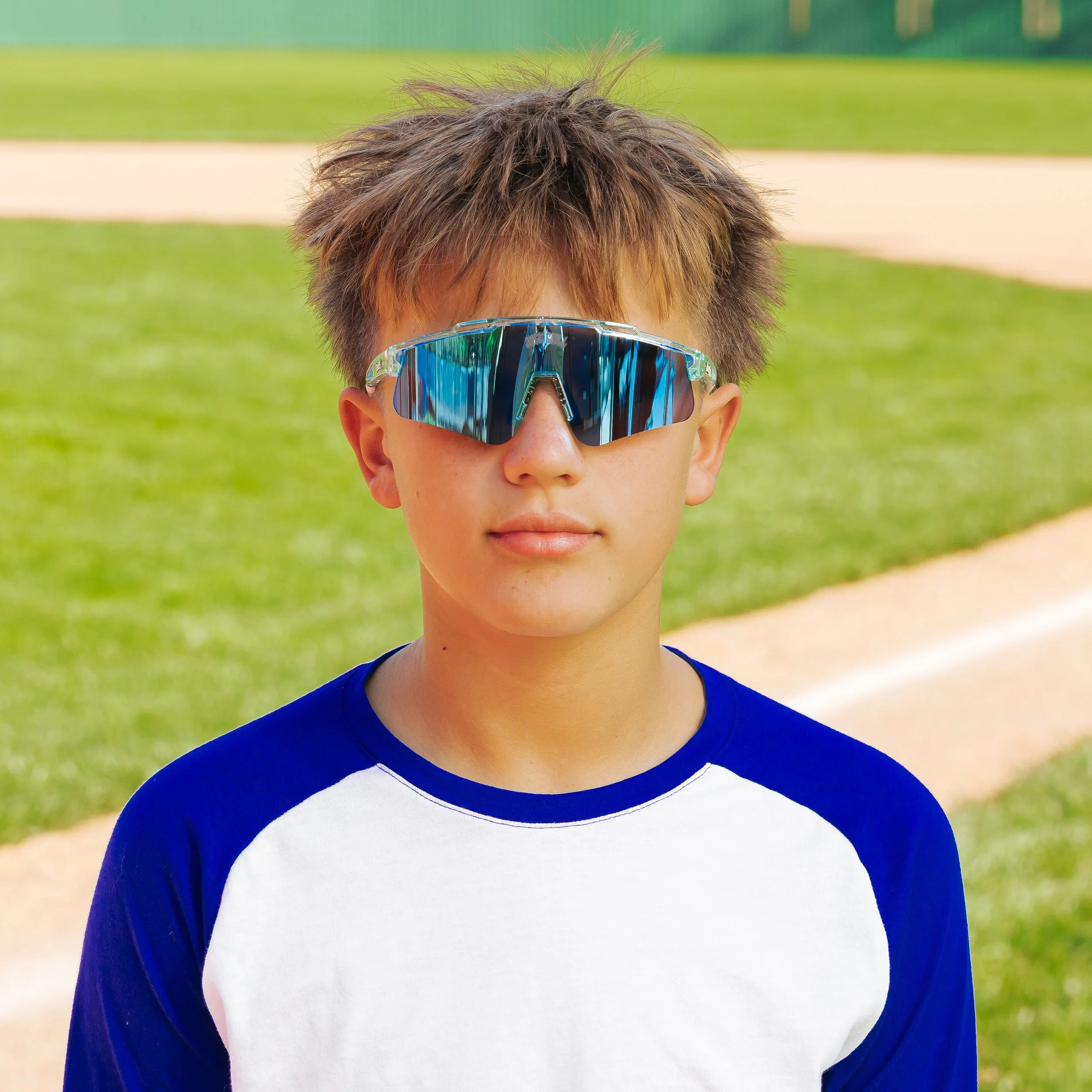 Close-up of a boy wearing sporty sunglasses and a blue and white baseball t-shirt, standing outdoors on a grassy field with a dirt path, during daytime.