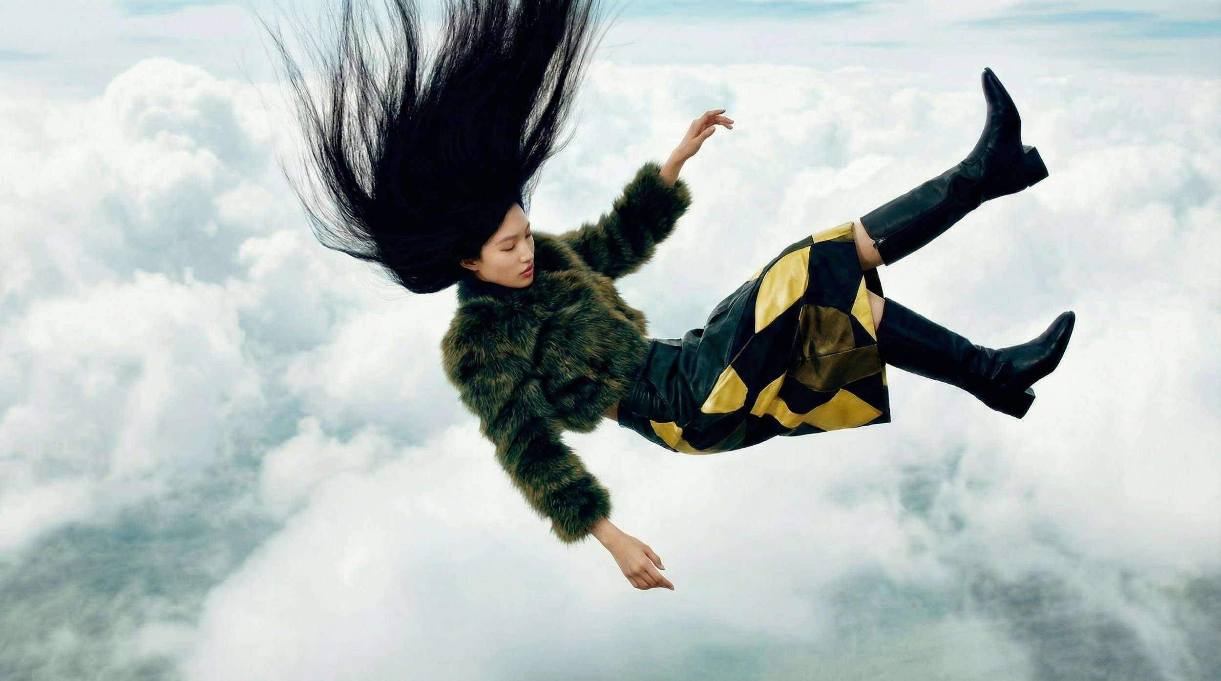 A woman with long black hair floating in the sky above clouds, wearing a fur coat and patterned pants.