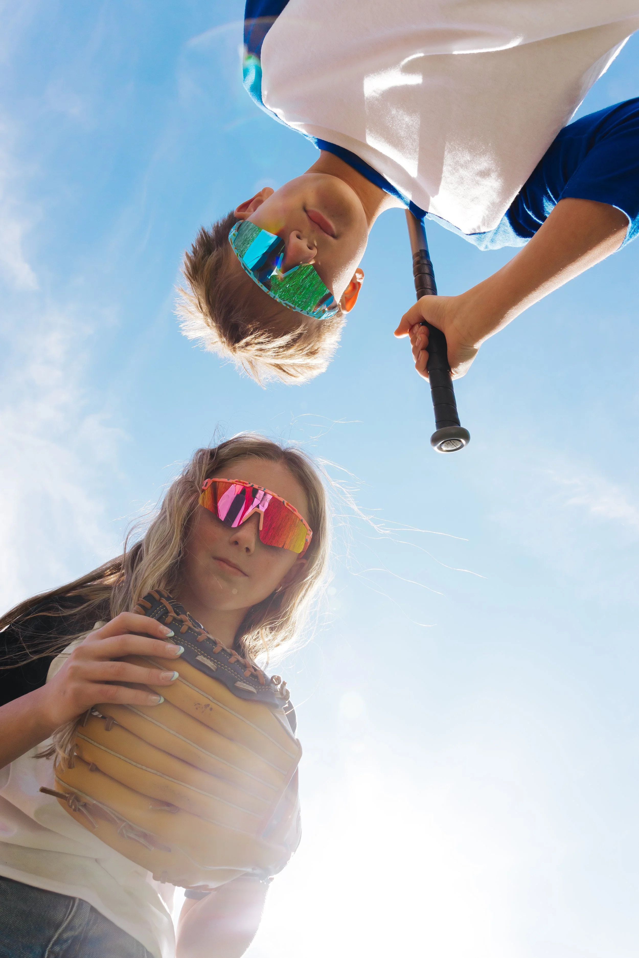 Young girl holding a baseball glove, wearing reflective sports sunglasses, with a woman in the background, bright sky and sunlight.