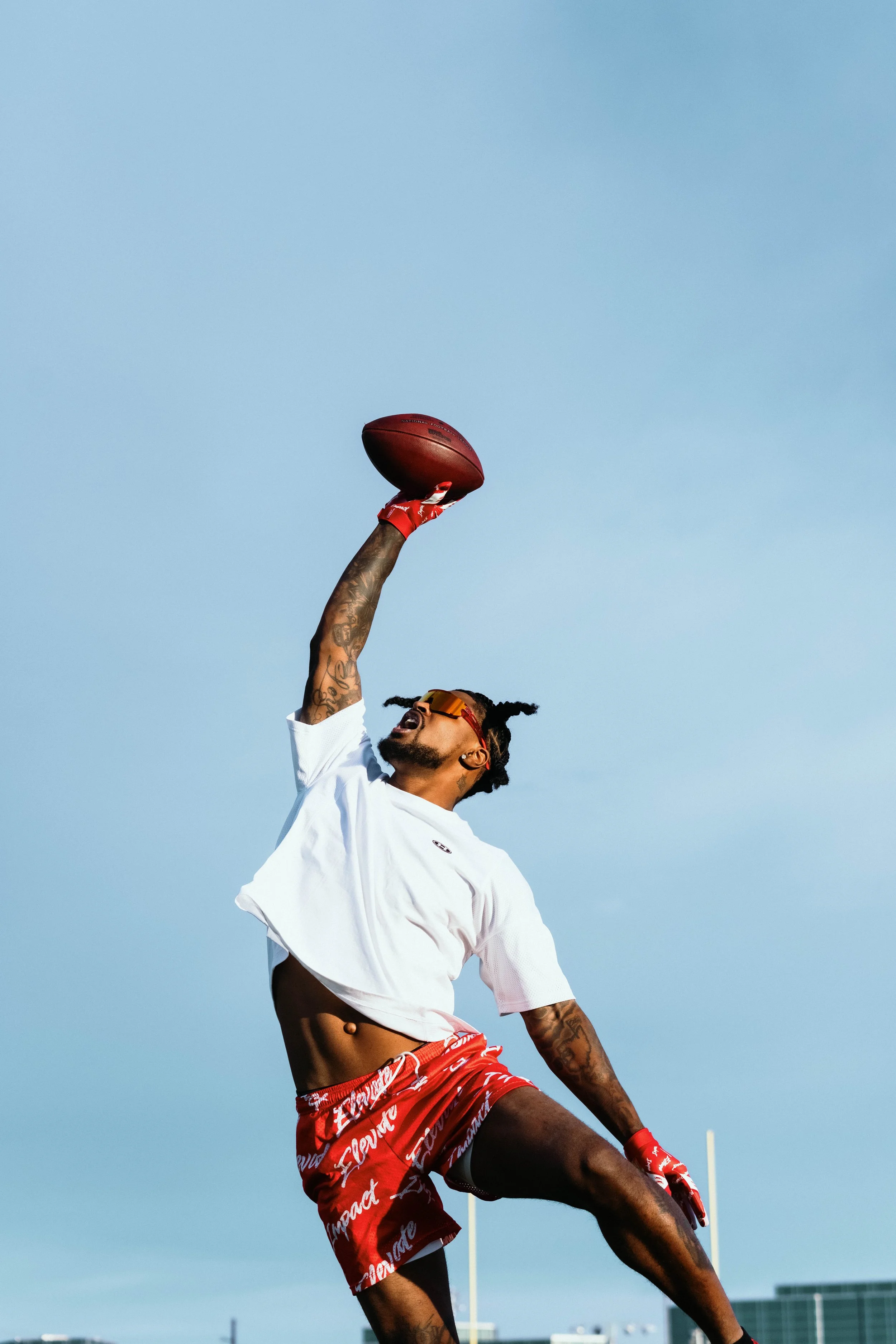 A man with tattoos on his arm is jumping in the air and holding a football above his head against a clear blue sky, wearing red shorts, sunglasses, and red gloves.