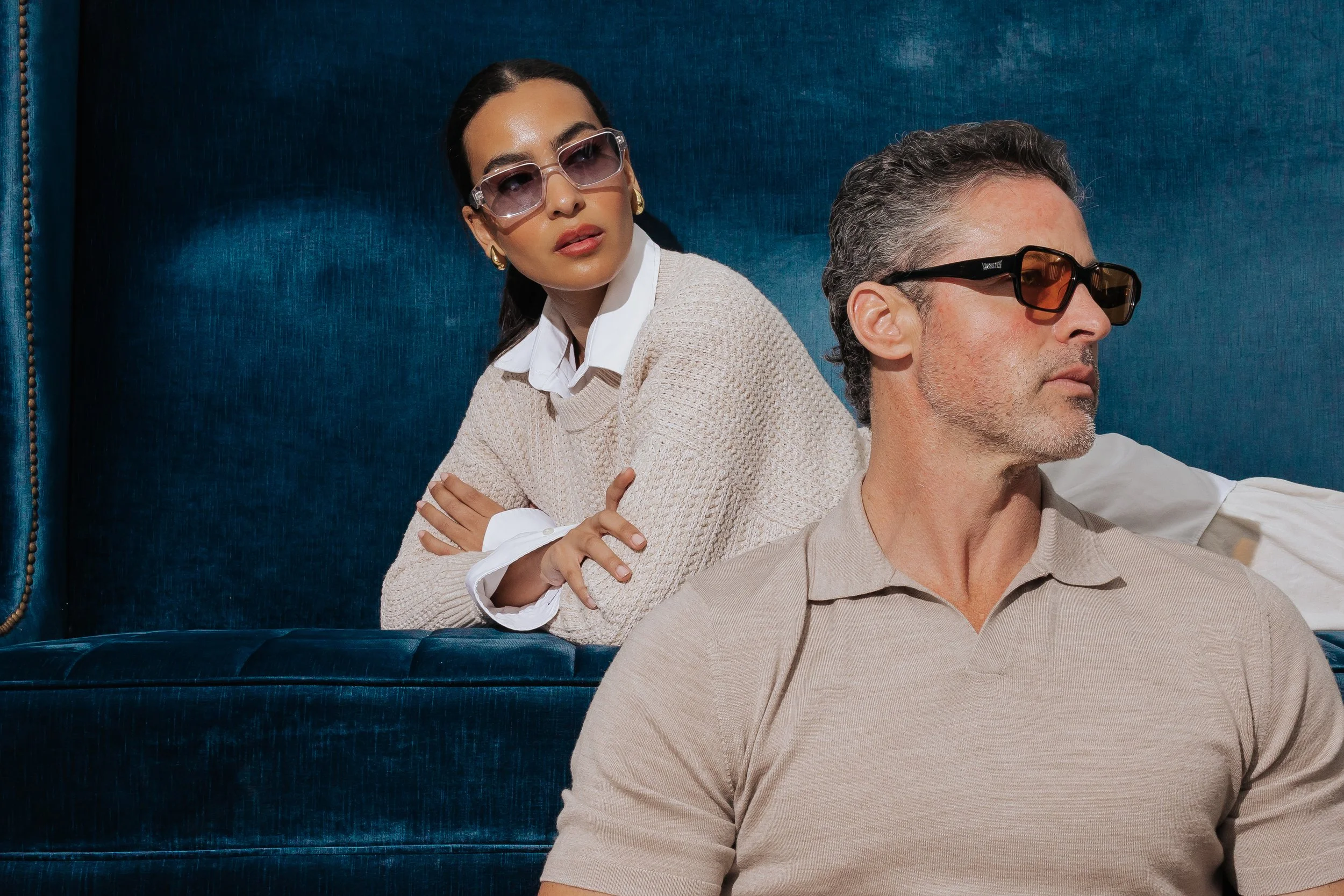 A woman with dark hair, wearing large sunglasses, a white collared shirt, and a beige sweater, is sitting with her arms crossed behind a man. The man has gray hair, is wearing sunglasses and a beige polo shirt, and is sitting in front of her. They are on a blue textured bench or headboard against a dark blue background.