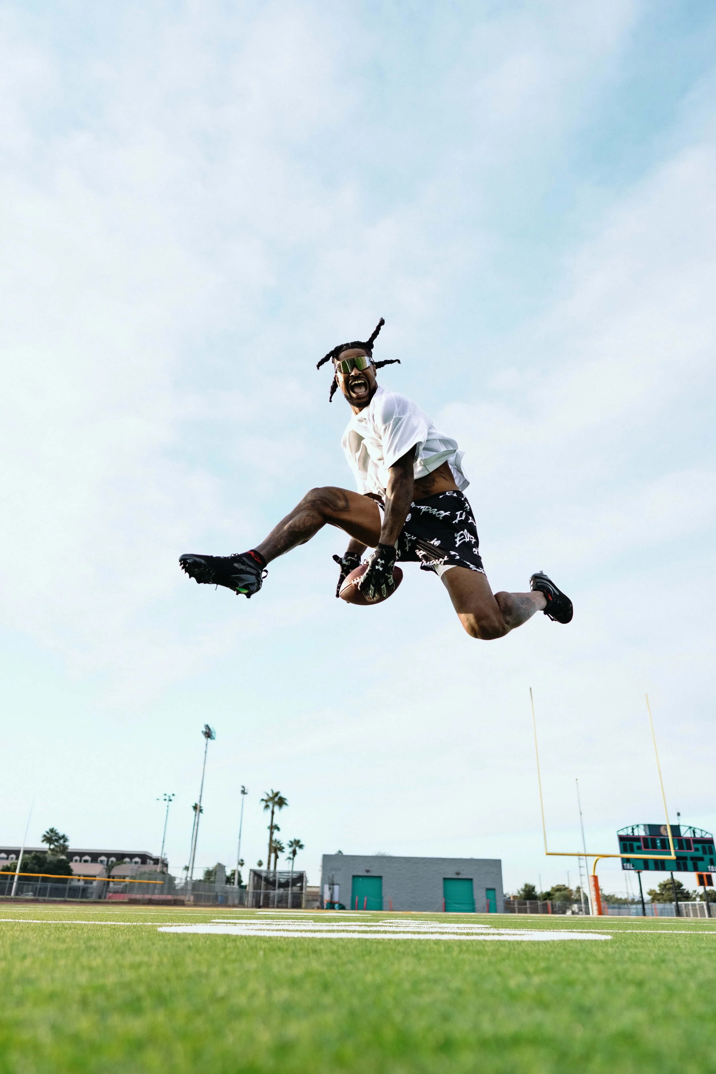A man performing a football stunt in a stadium, mid-air, with the sky and stadium lights in the background. He appears energetic and intense, dressed in athletic gear, with a football.