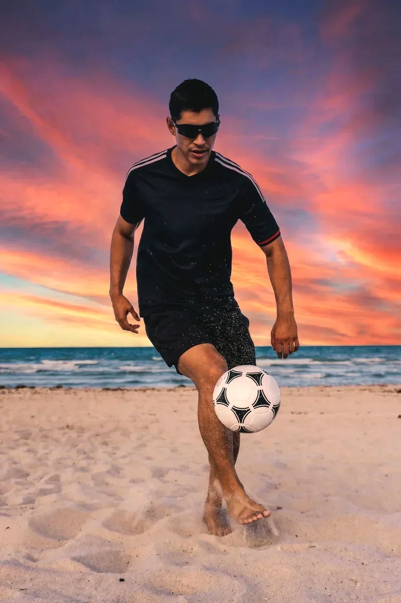 Man wearing sunglasses playing soccer on the beach at sunset.