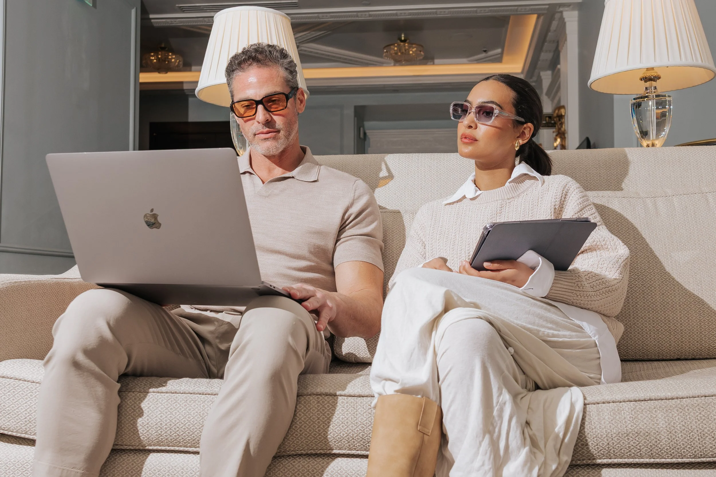 A middle-aged man and a woman sitting on a beige sofa in a luxurious, well-lit living room, both wearing glasses and focused on devices; the man has a laptop on his lap, and the woman is holding a tablet.