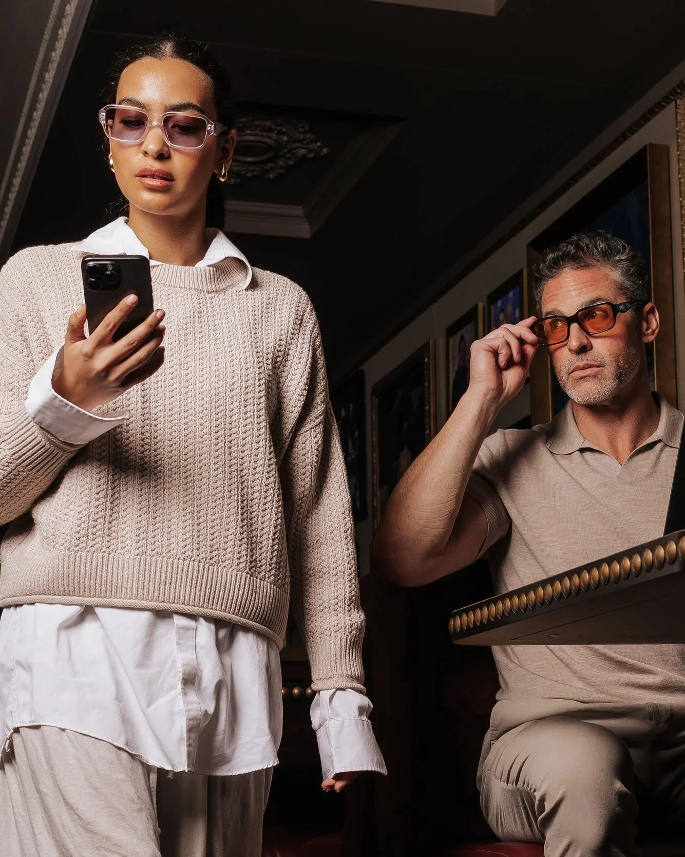 A woman in glasses and a beige sweater looks at her phone, while a man in glasses looks at a framed mirror, in an indoor setting, possibly a gallery.