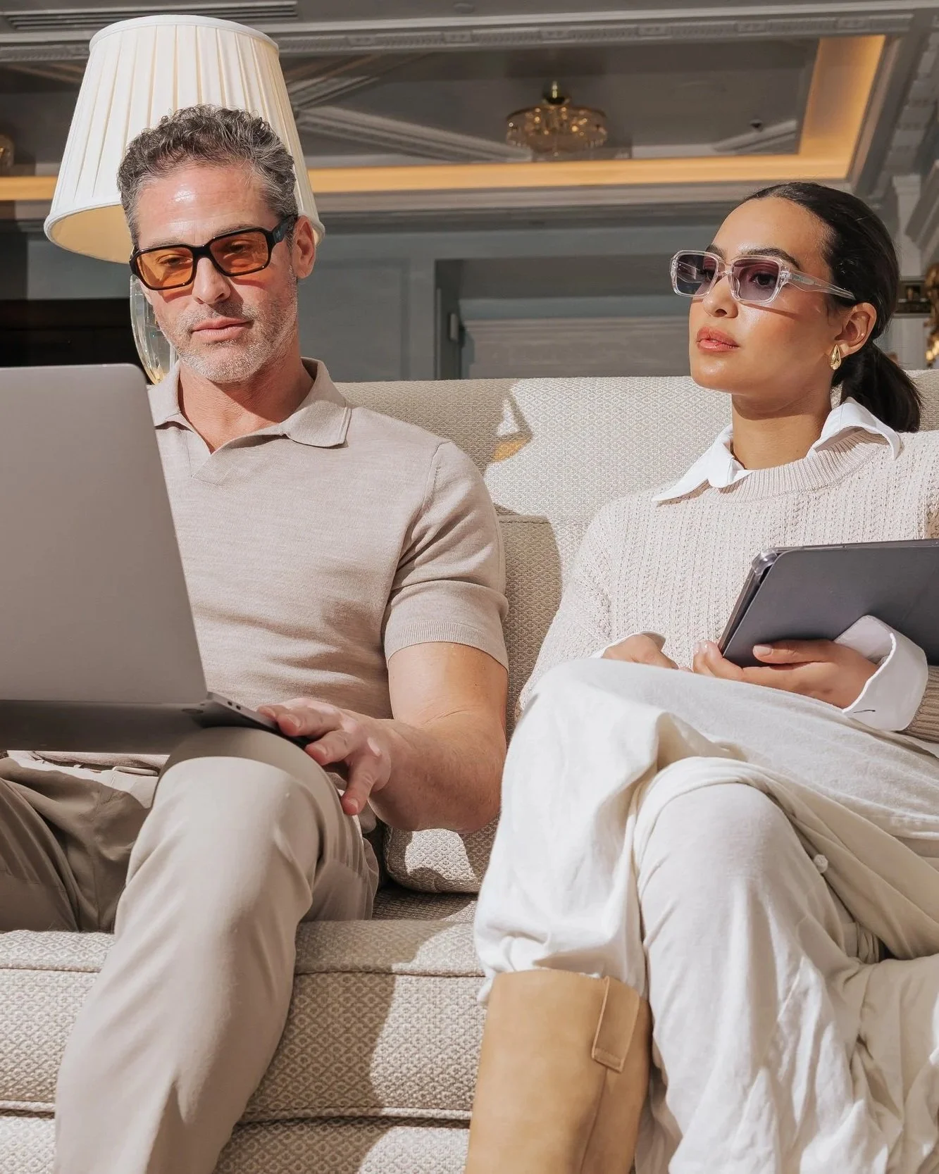 A man and woman sit on a beige sofa in a luxurious room, both wearing glasses. The man is using a laptop, while the woman holds a tablet.