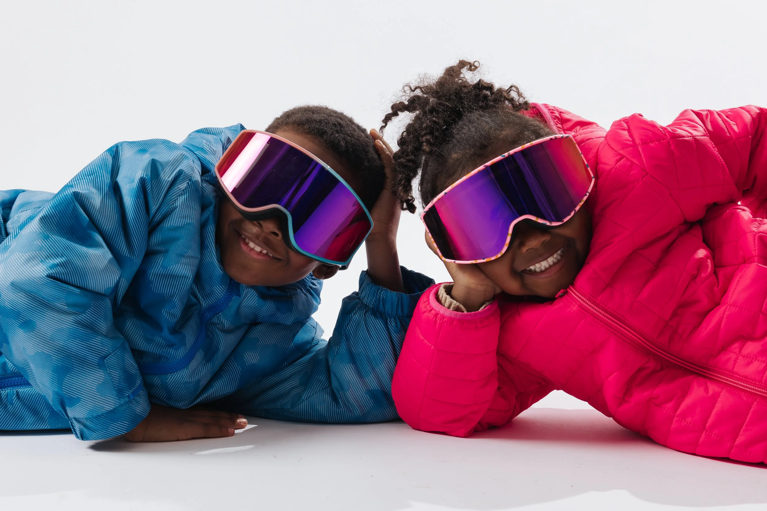 Two smiling children, one wearing a blue jacket and the other a pink jacket, lying on the ground and touching foreheads, wearing large colorful sunglasses.