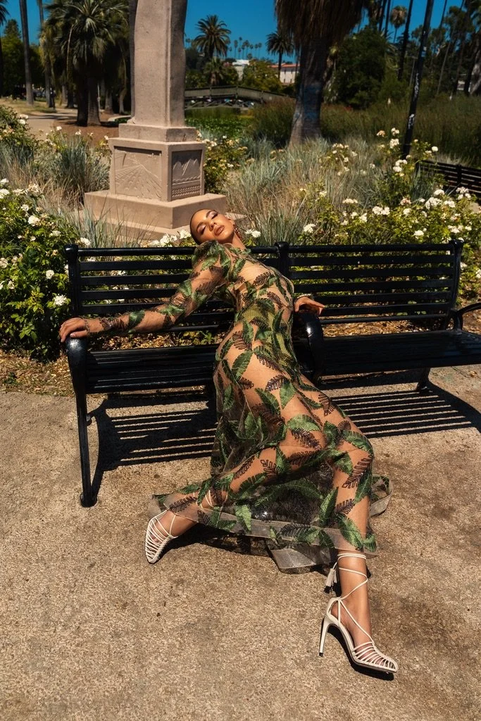 A woman in a sheer dress with green leaf patterns is posing on a park bench with her arms outstretched, head tilted back, and eyes closed, under clear blue skies with palm trees and greenery in the background.