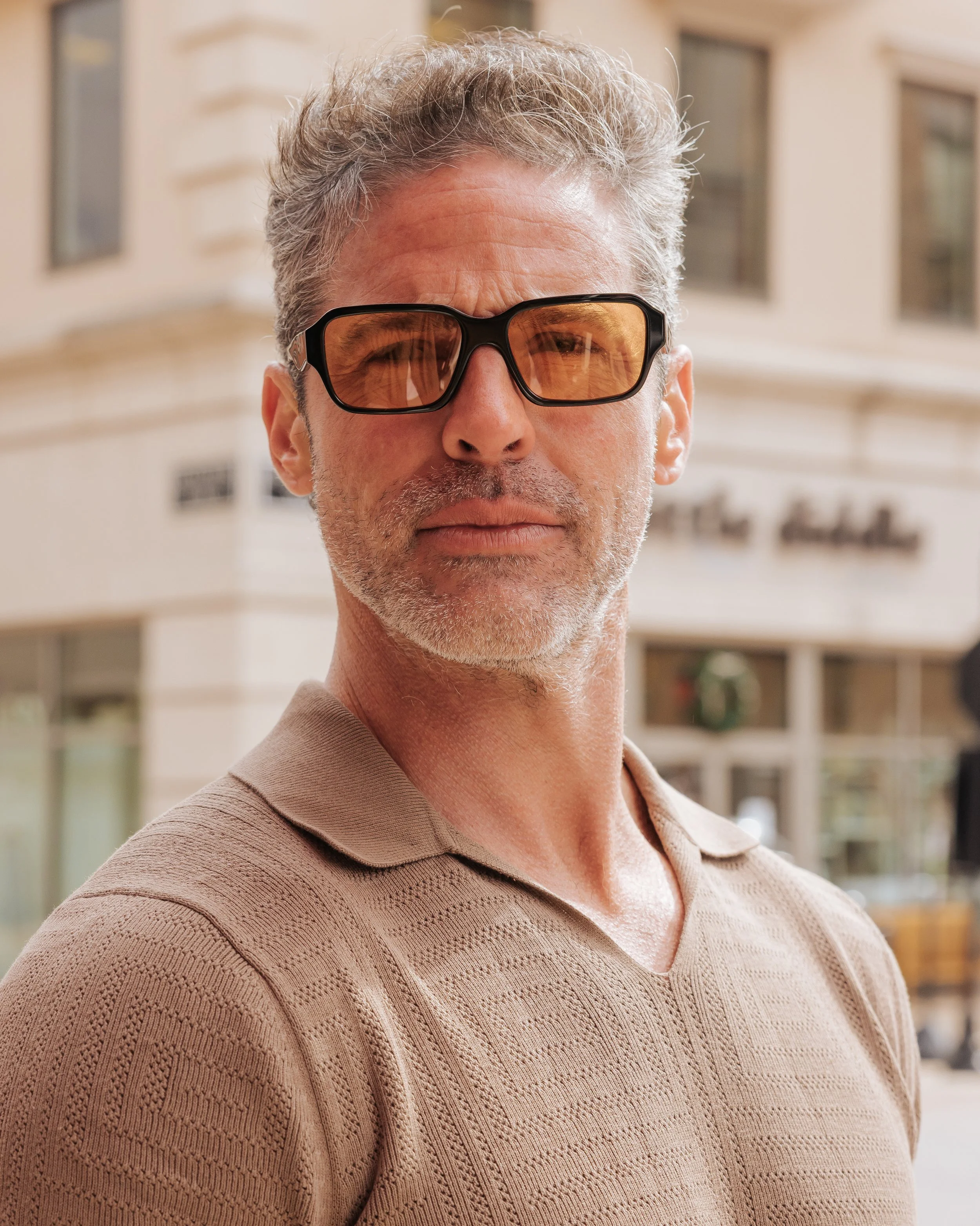 Close-up of a middle-aged man with gray hair and stubble wearing sunglasses and a beige knitted polo shirt, outdoors in front of a blurred building.
