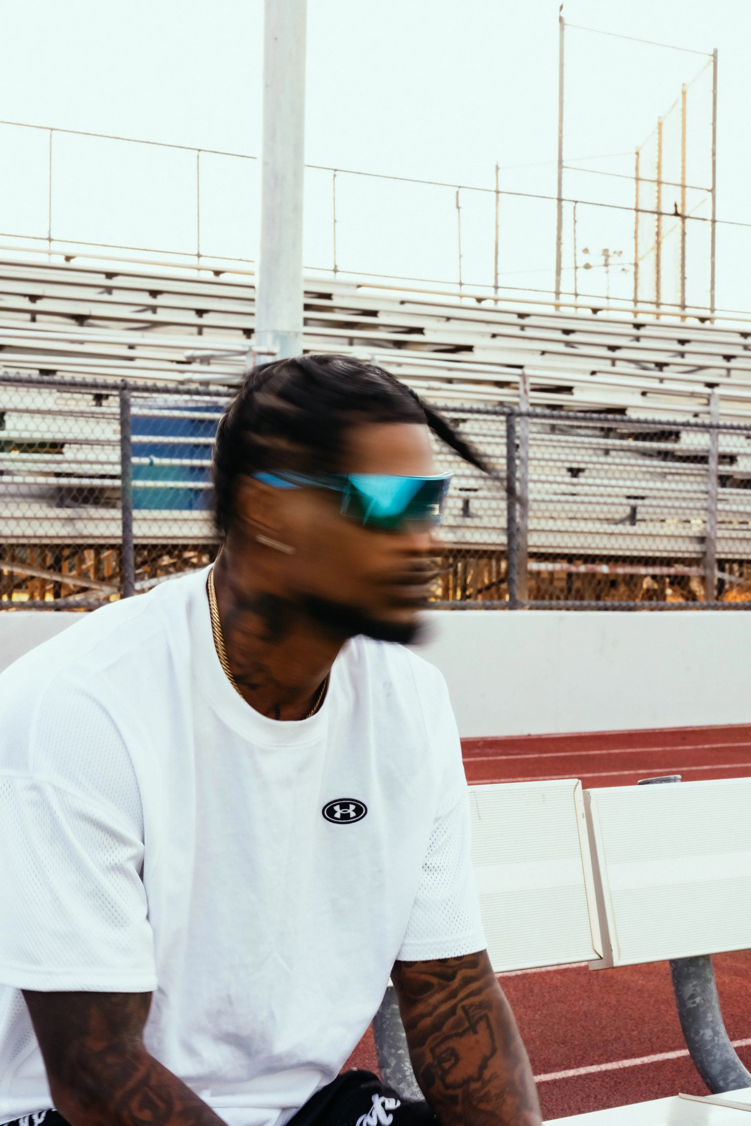A man with tattoos on his arms wearing sunglasses and a white Under Armour shirt is sitting outdoors near a track and sports bleachers.